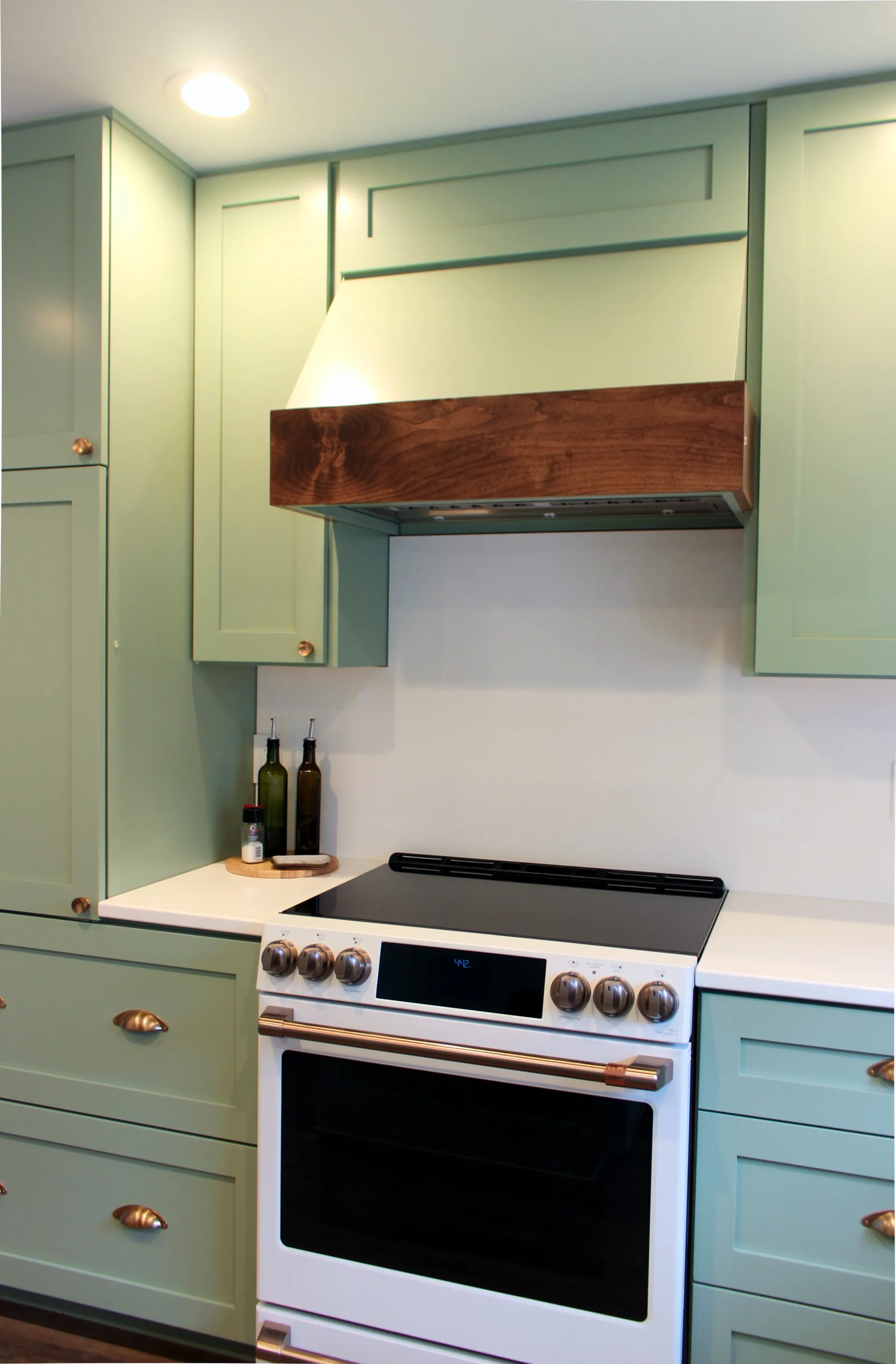 A photograph of custom light green kitchen cabinets, a café range/oven, and a custom vent hood with wood detail.