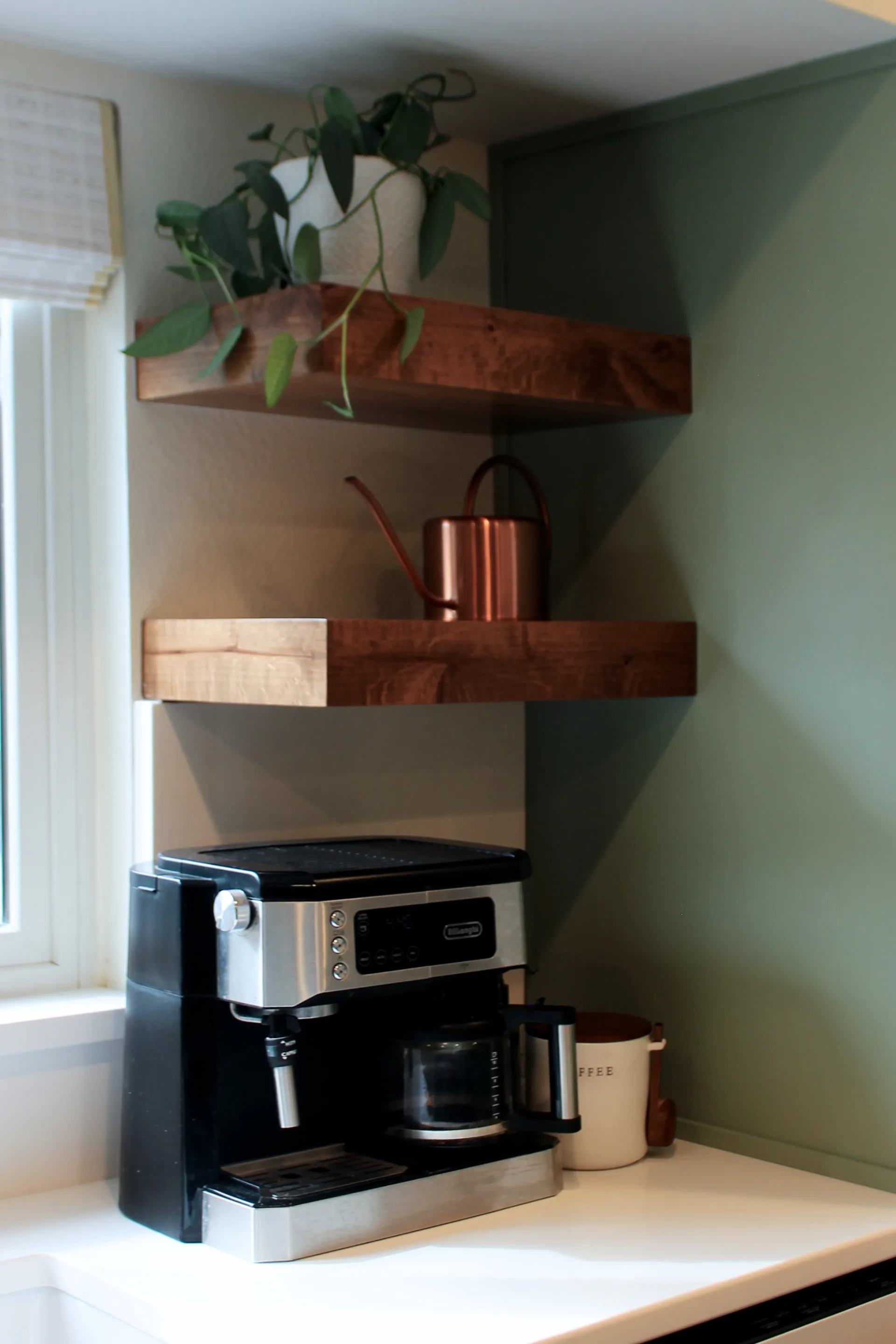 A close up photograph of custom wood floating shelves with an espresso maker, a copper watering can, and a plant.