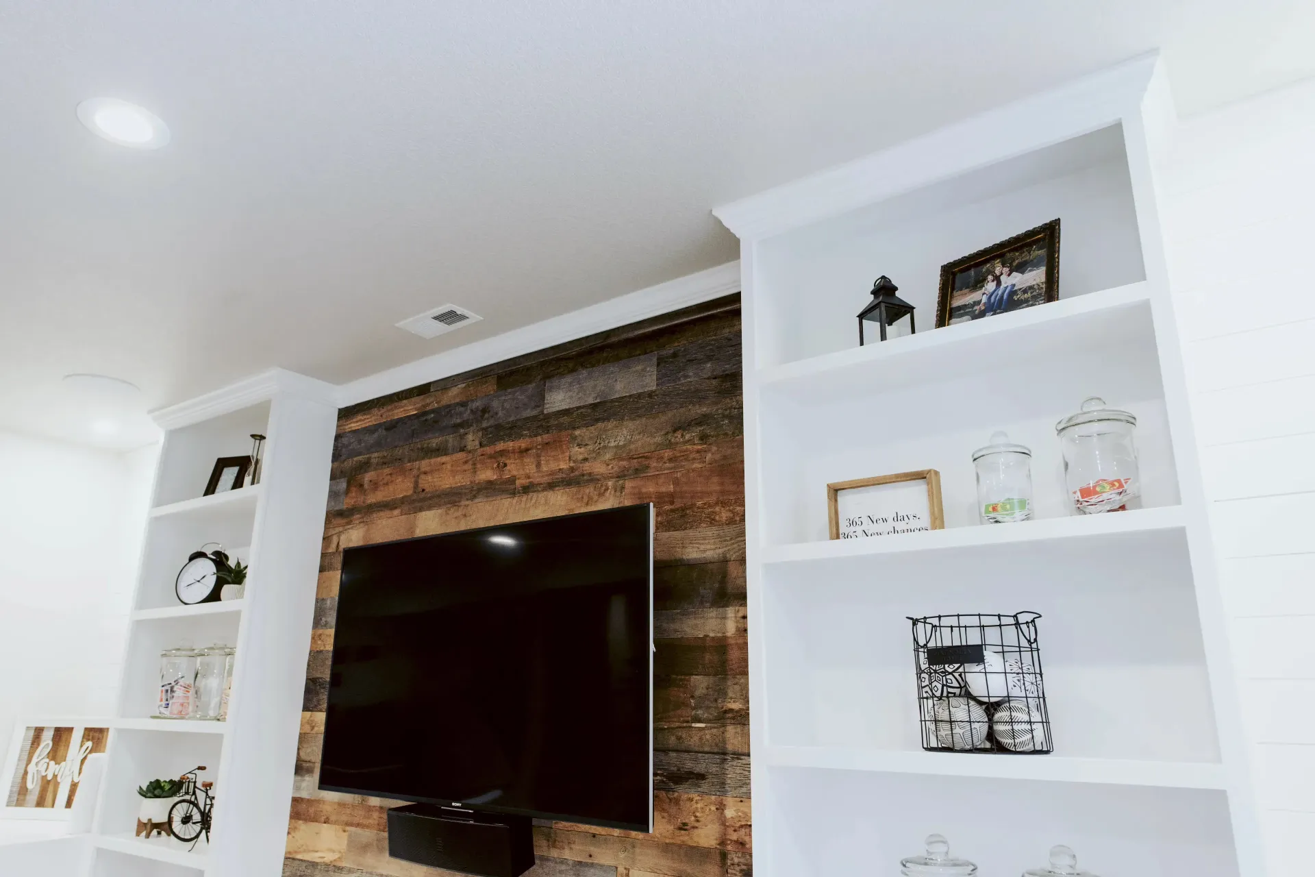 Living room with a large flat-screen TV mounted on a rustic wooden wall. Flanked by white shelves holding jars, baskets, and framed photos. Cozy and modern ambiance.