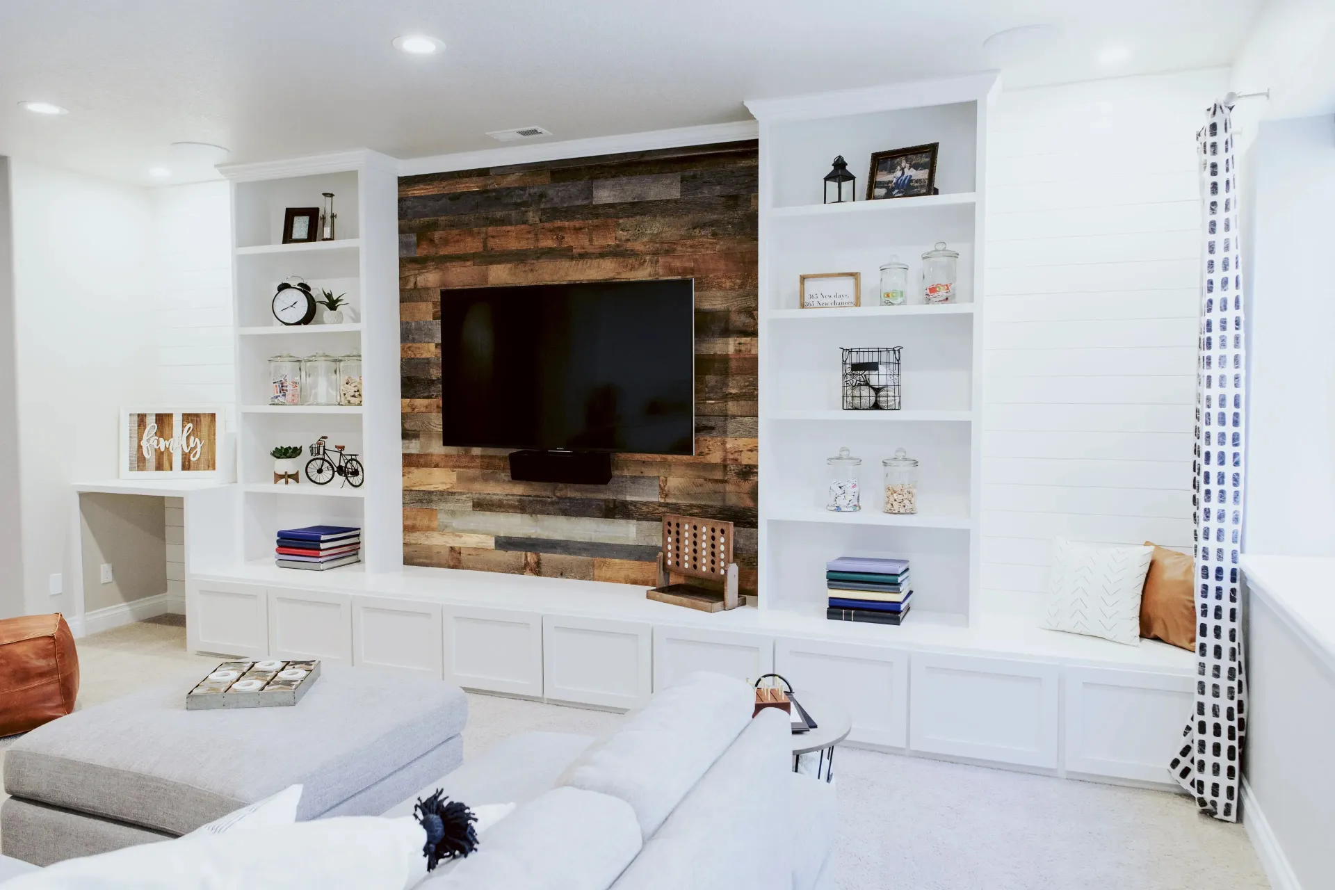 Modern living room with a wall-mounted TV on wooden accent wall, flanked by white shelves holding books, plants, and decor. Neutral tones create a cozy atmosphere.