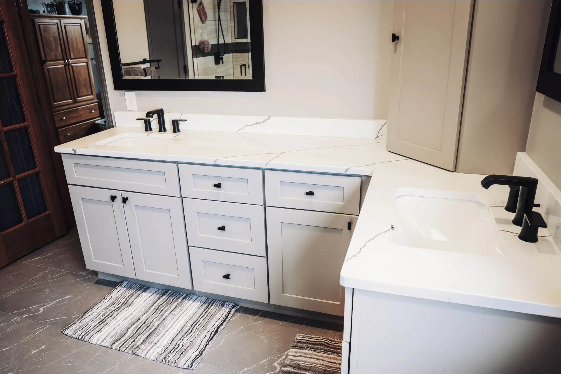 Modern bathroom with light gray double-sink vanity, black fixtures, marble countertop, striped rugs, large mirror, and a wooden cabinet in the background.