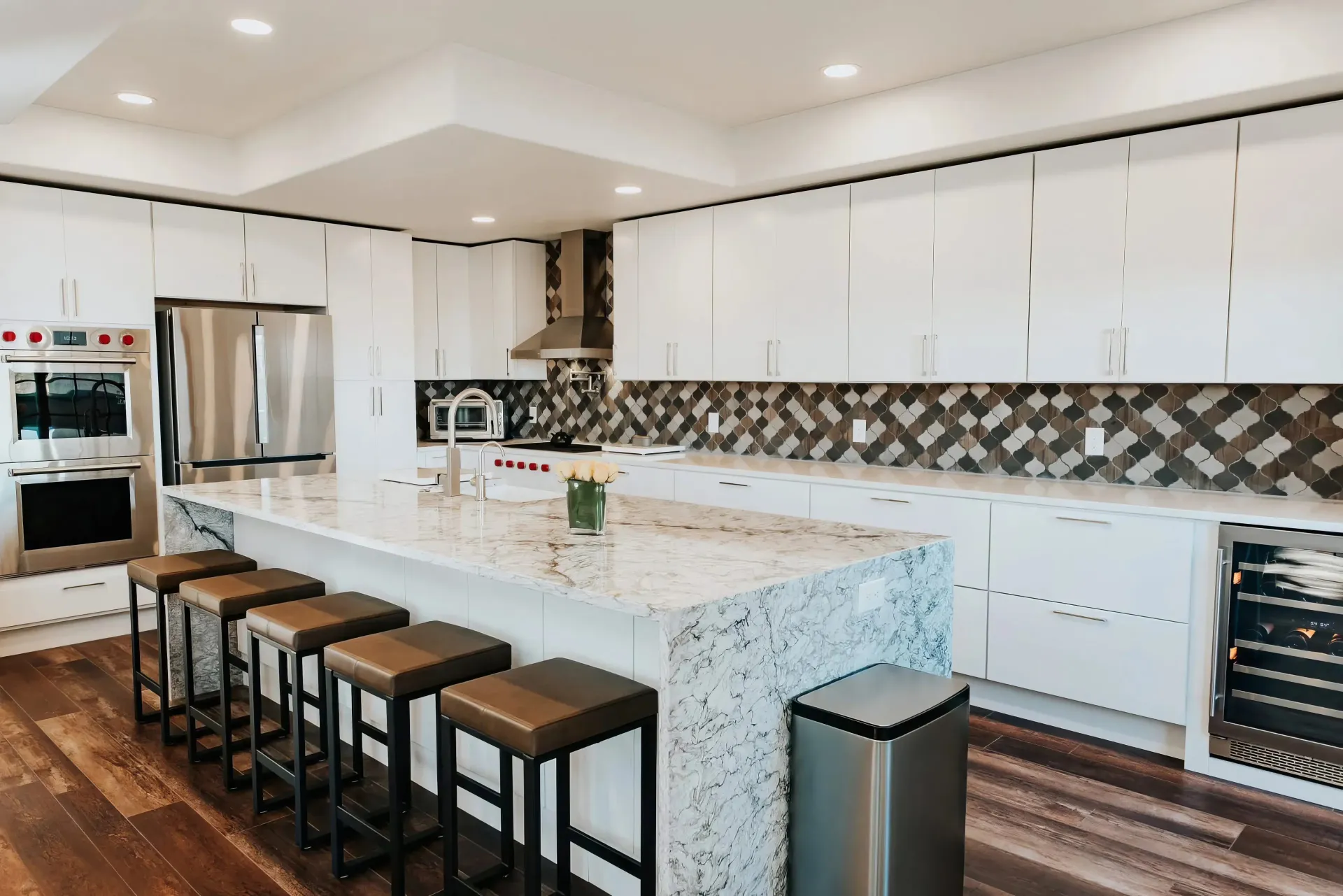 Modern kitchen featuring white cabinets, a marble island with five cushioned stools, stainless steel appliances, and geometric backsplash. Warm, inviting tone.