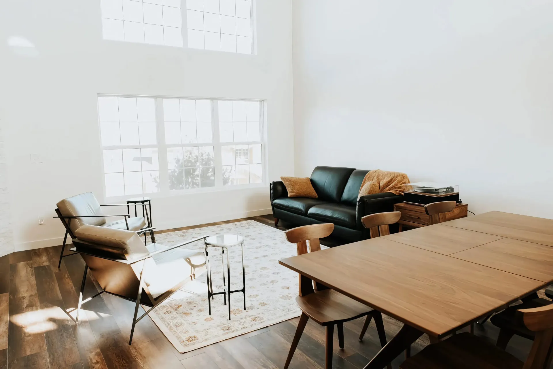 Modern living room with high ceilings, large windows, and sunlight. Features a black leather sofa, chairs, dining table, and cozy decor with wooden accents.