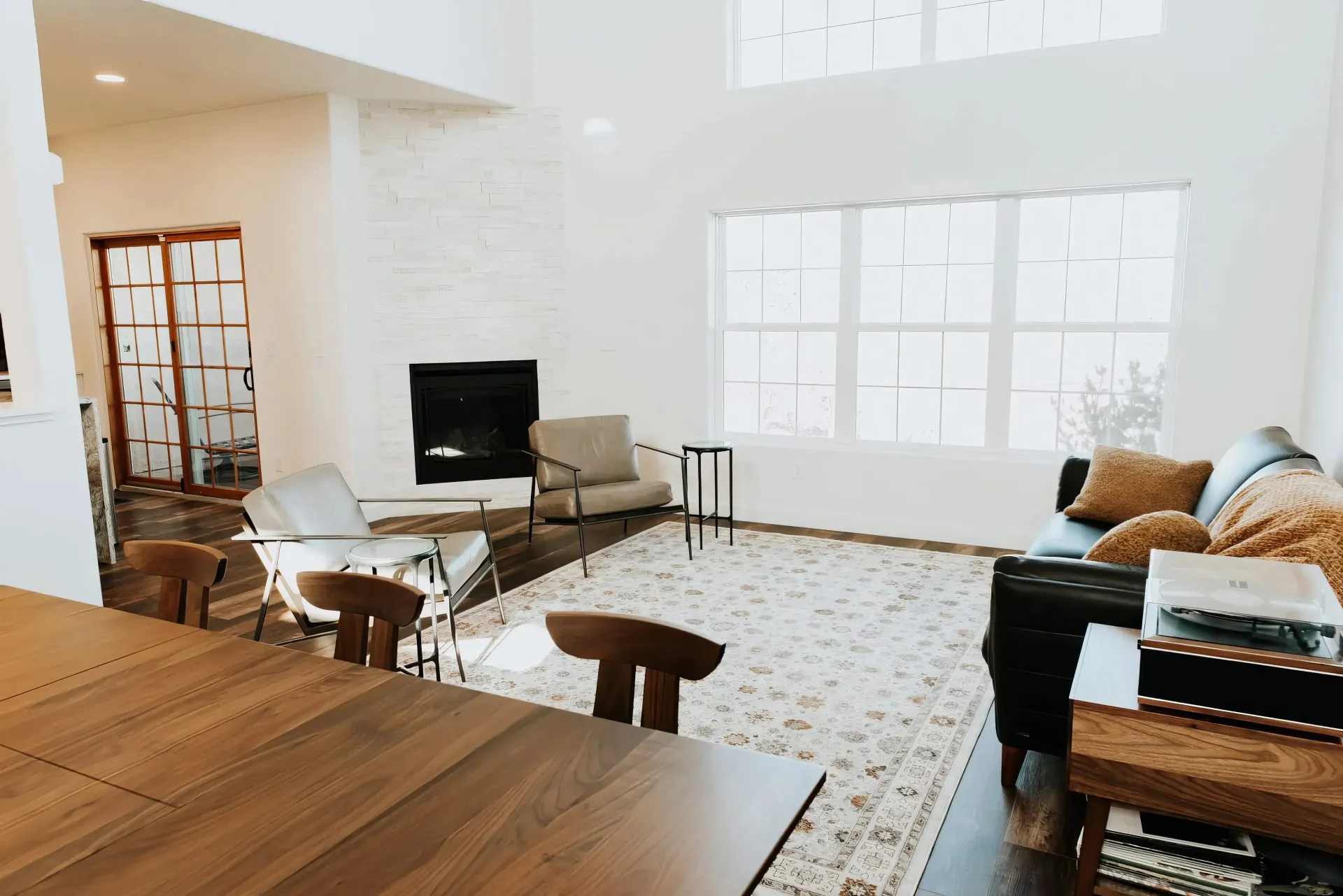 Stylish living room with a dark leather sofa, cushioned chairs, and a modern fireplace. Sunlight streams through large windows, highlighting the area rug and wooden furniture, creating a warm and inviting atmosphere.