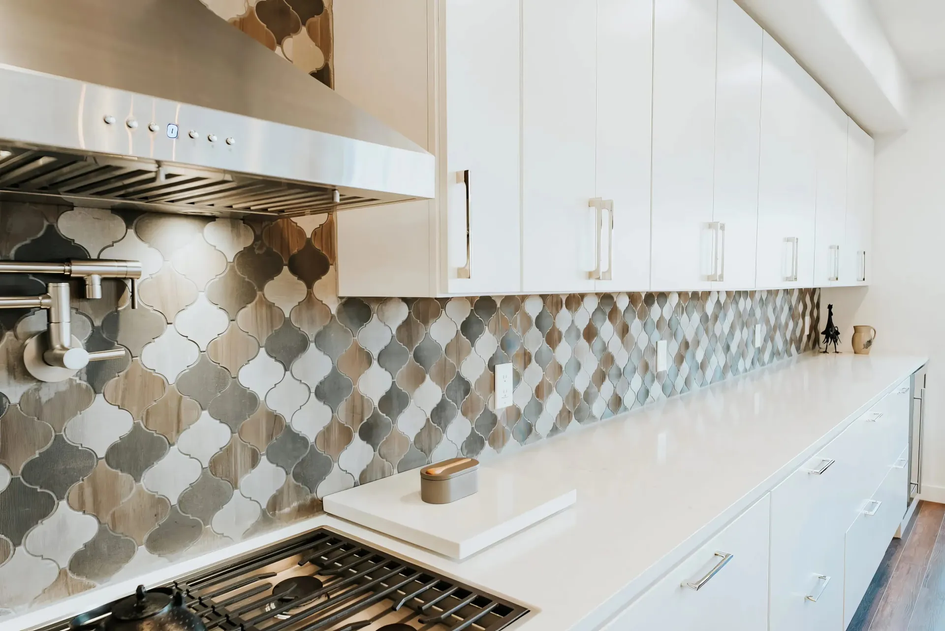 Modern kitchen with a white countertop, geometric tile backsplash, and sleek white cabinets. A stainless steel range hood and stove add elegance.