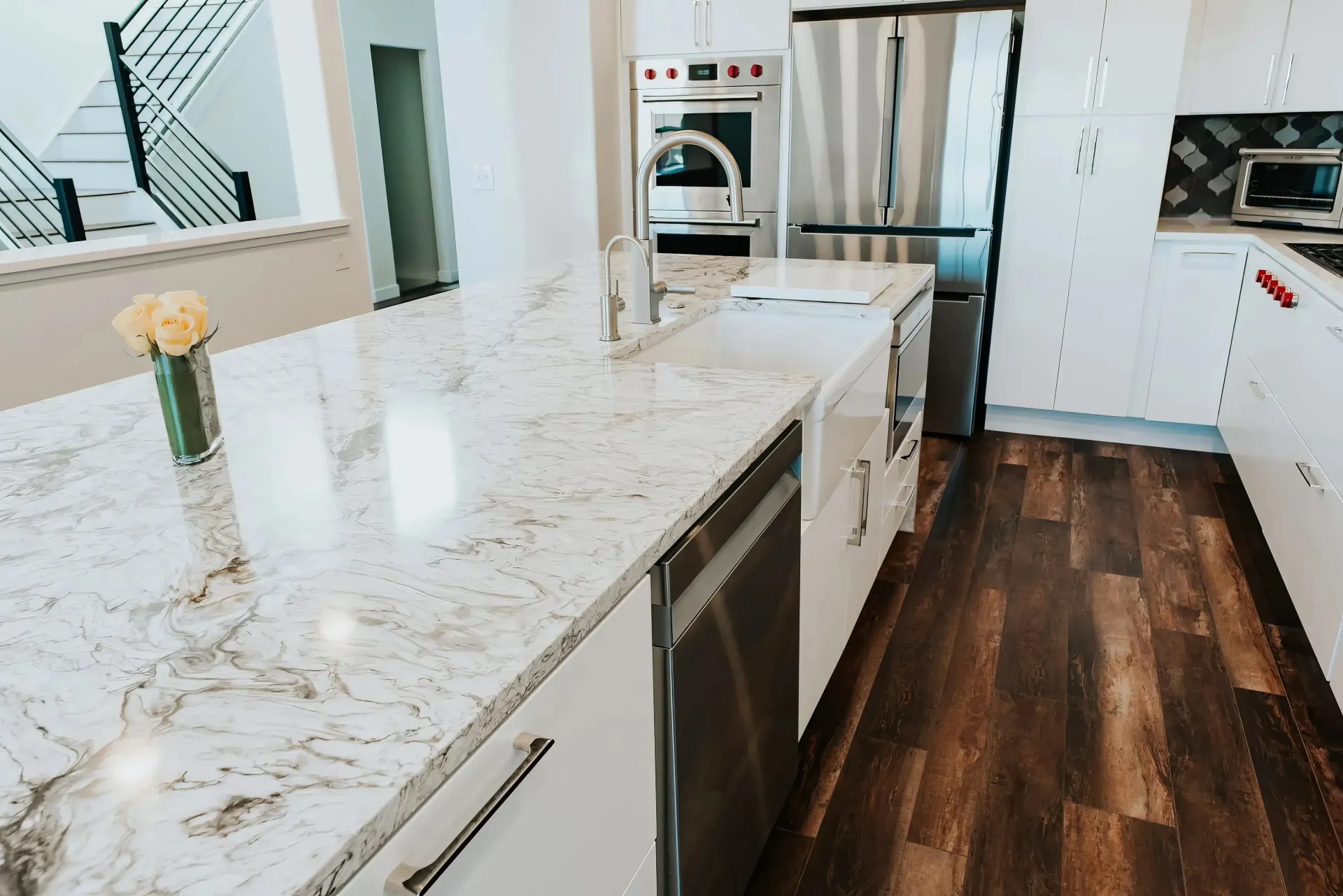 Modern kitchen with a marble island, stainless steel appliances, and wood flooring. Vase with yellow roses adds a touch of elegance. Bright and inviting.