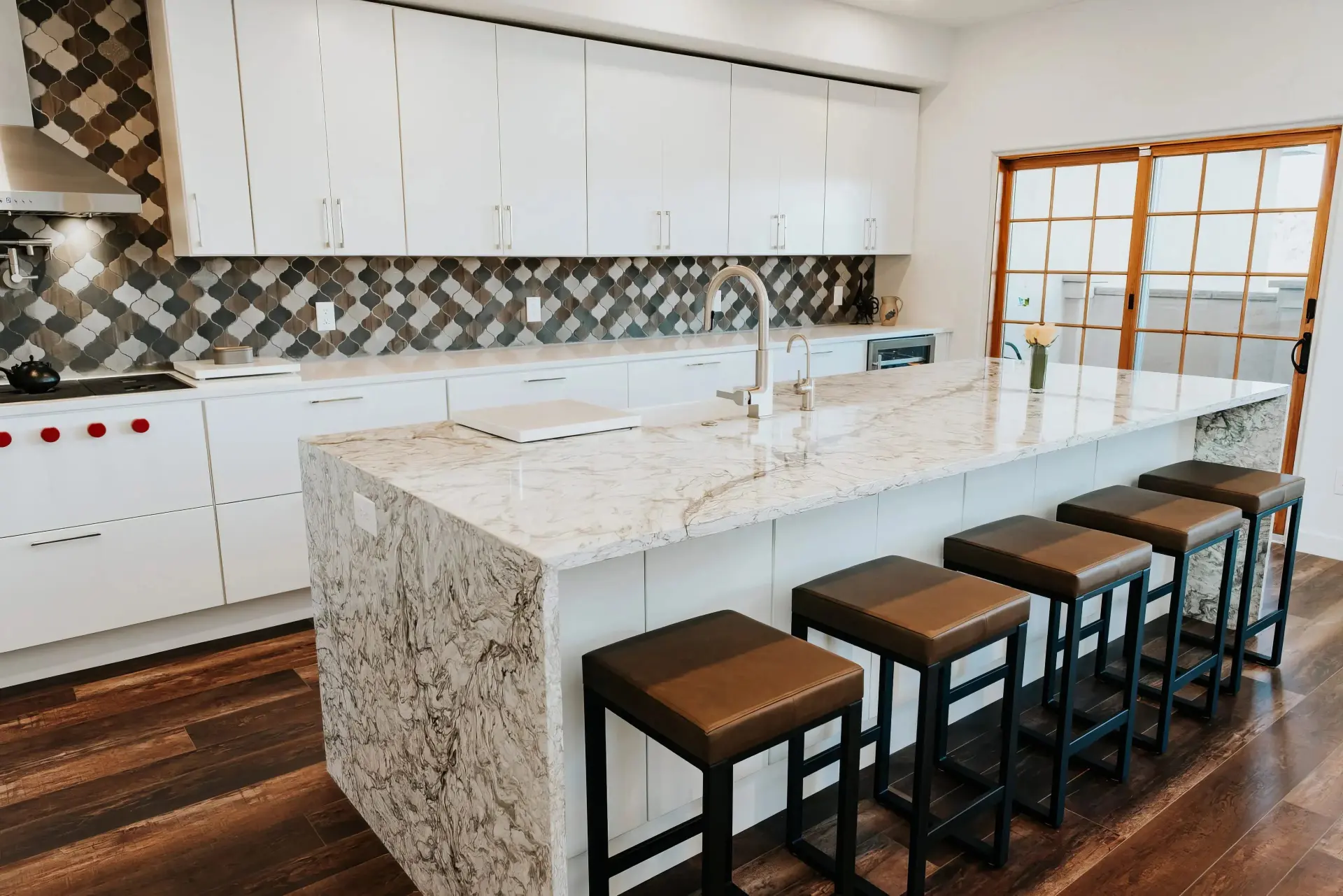 Modern kitchen with a marble island, sleek white cabinets, and a geometric tiled backsplash. Five stools line the island. Warm, inviting ambiance.