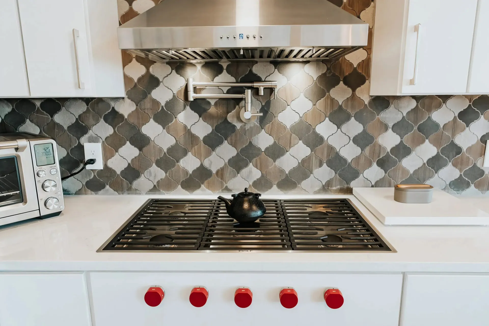 Modern kitchen with geometric tile backsplash, stainless hood, and stove. A black kettle and red-knobbed controls add contrast. Bright, clean ambiance.
