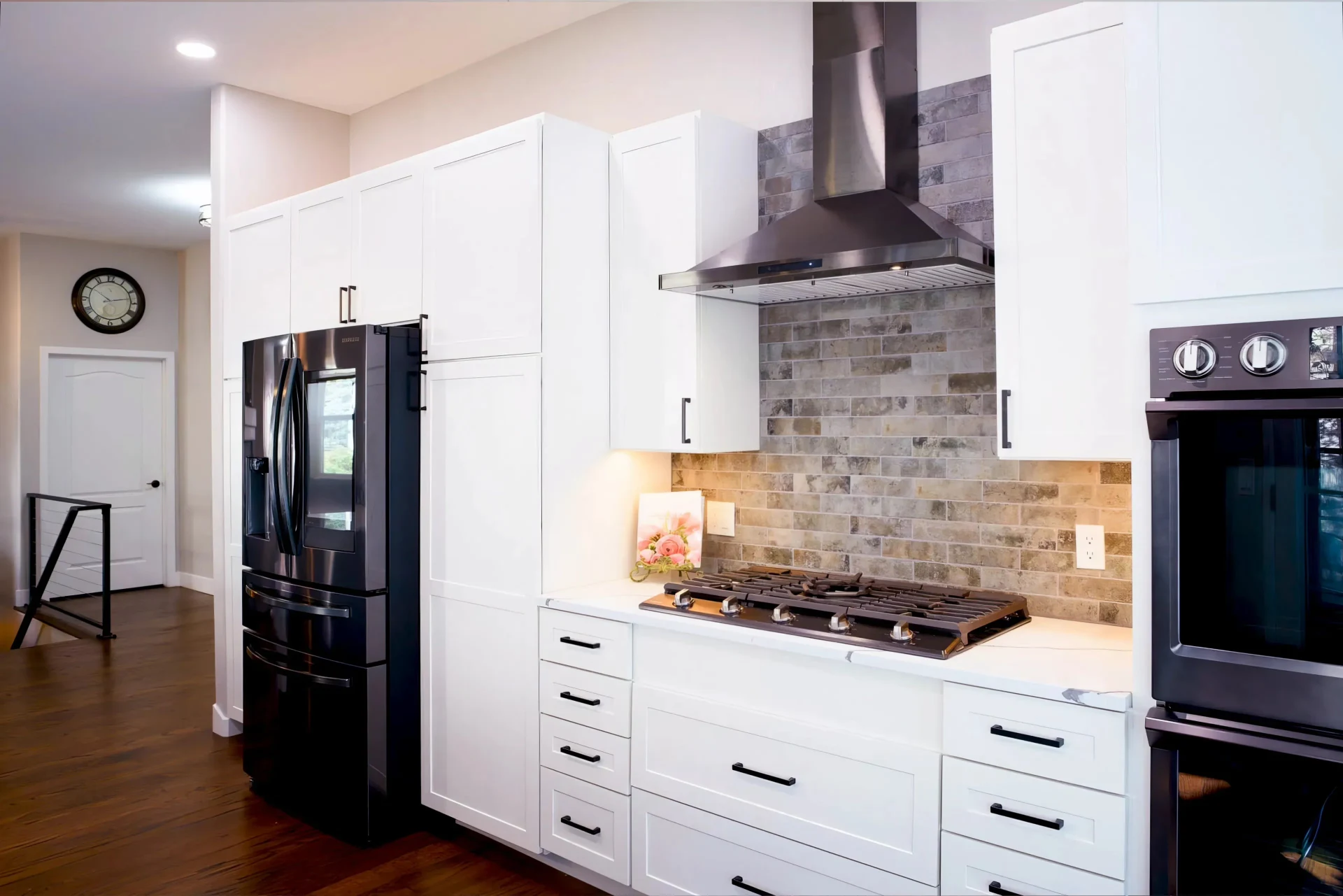 Modern kitchen with white cabinets, black appliances, brick backsplash, and stovetop. Warm lighting and hardwood floors create a cozy atmosphere.