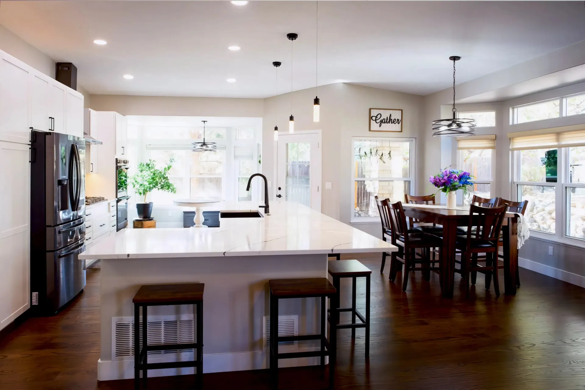 Bright kitchen and dining area with white cabinets, marble countertops, and dark wood floors. A round table with a vase of purple flowers sits near large windows, under a sign reading "Gather." Modern pendant lights hang overhead, creating a warm, inviting