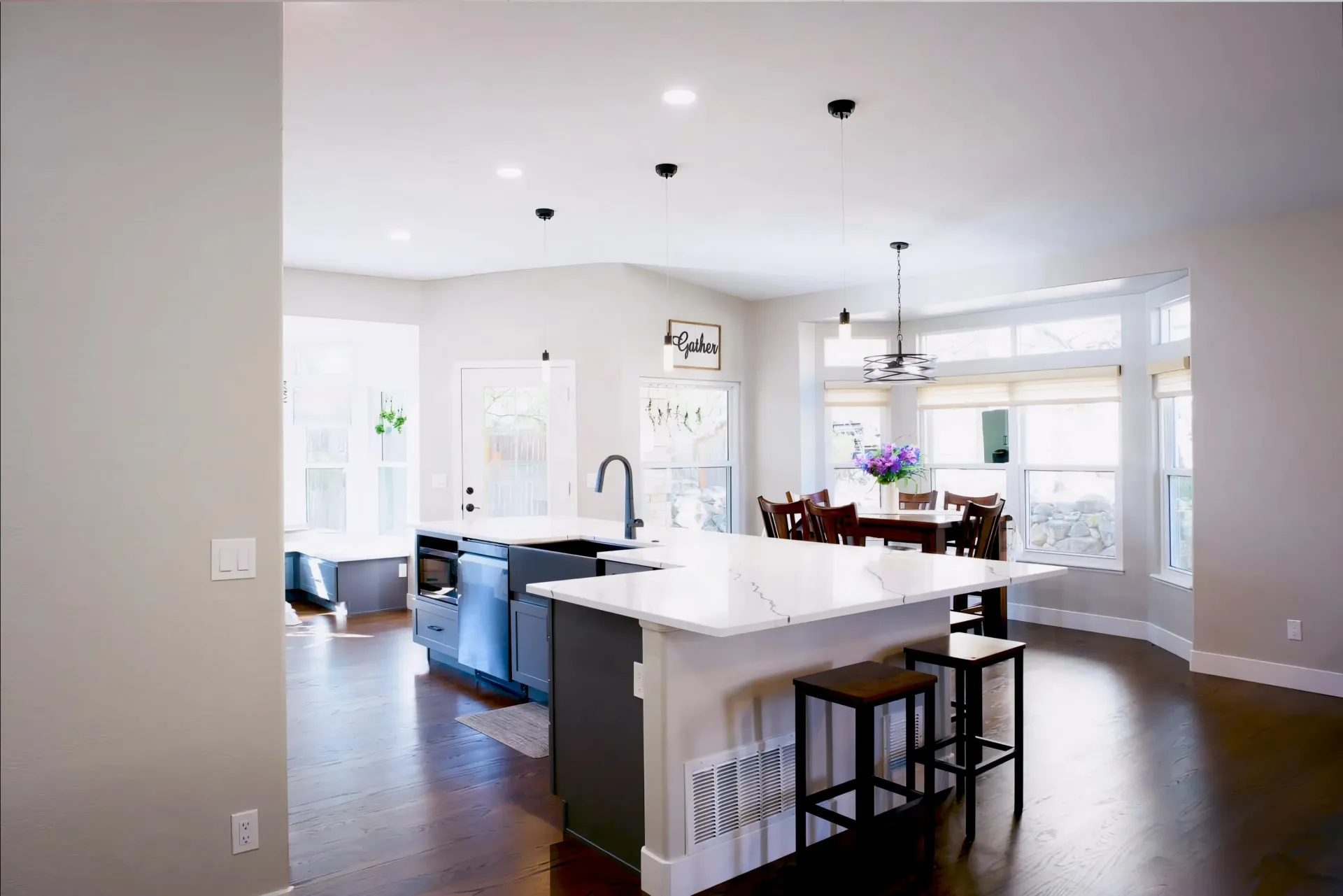 Modern kitchen and dining area with white countertops and pendant lights. Wooden floors, large windows, and purple flowers create a bright, welcoming space.