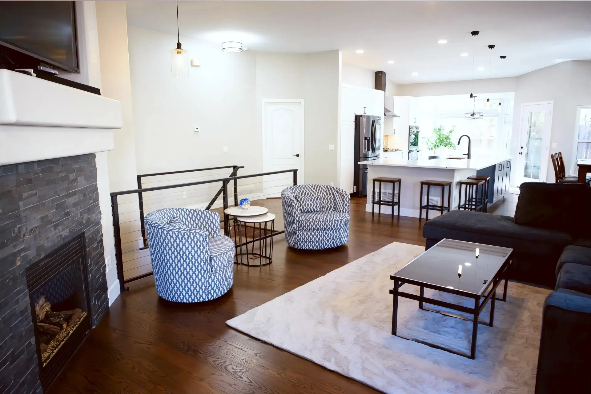 Modern open-concept living room and kitchen with a cozy ambiance. Features a stone fireplace, patterned armchairs, a plush rug, and a bright kitchen island.