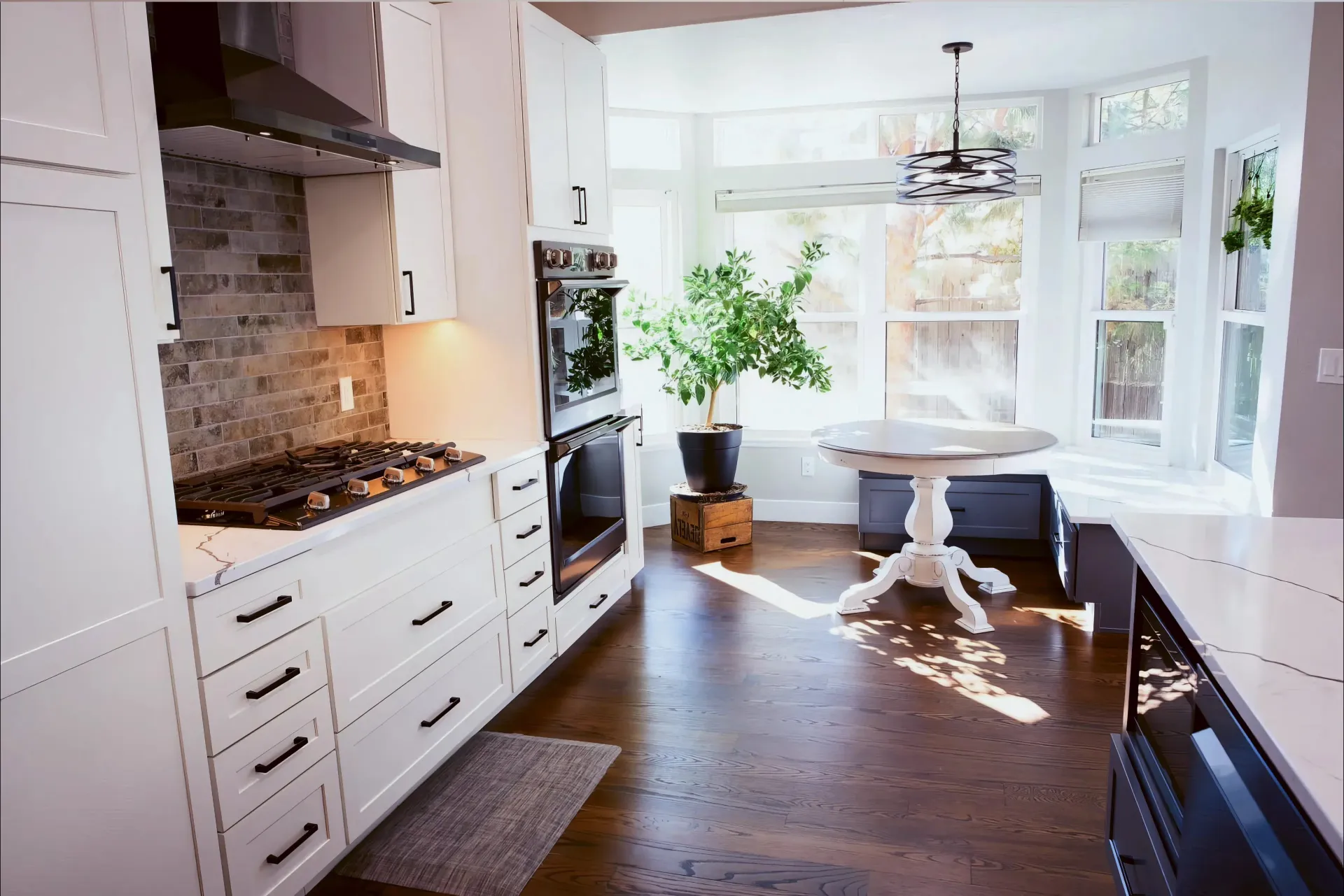 Bright kitchen with white cabinets, black accents, a brick backsplash, and stainless steel appliances. Sunlit dining nook with a round table and potted plant adds warmth.