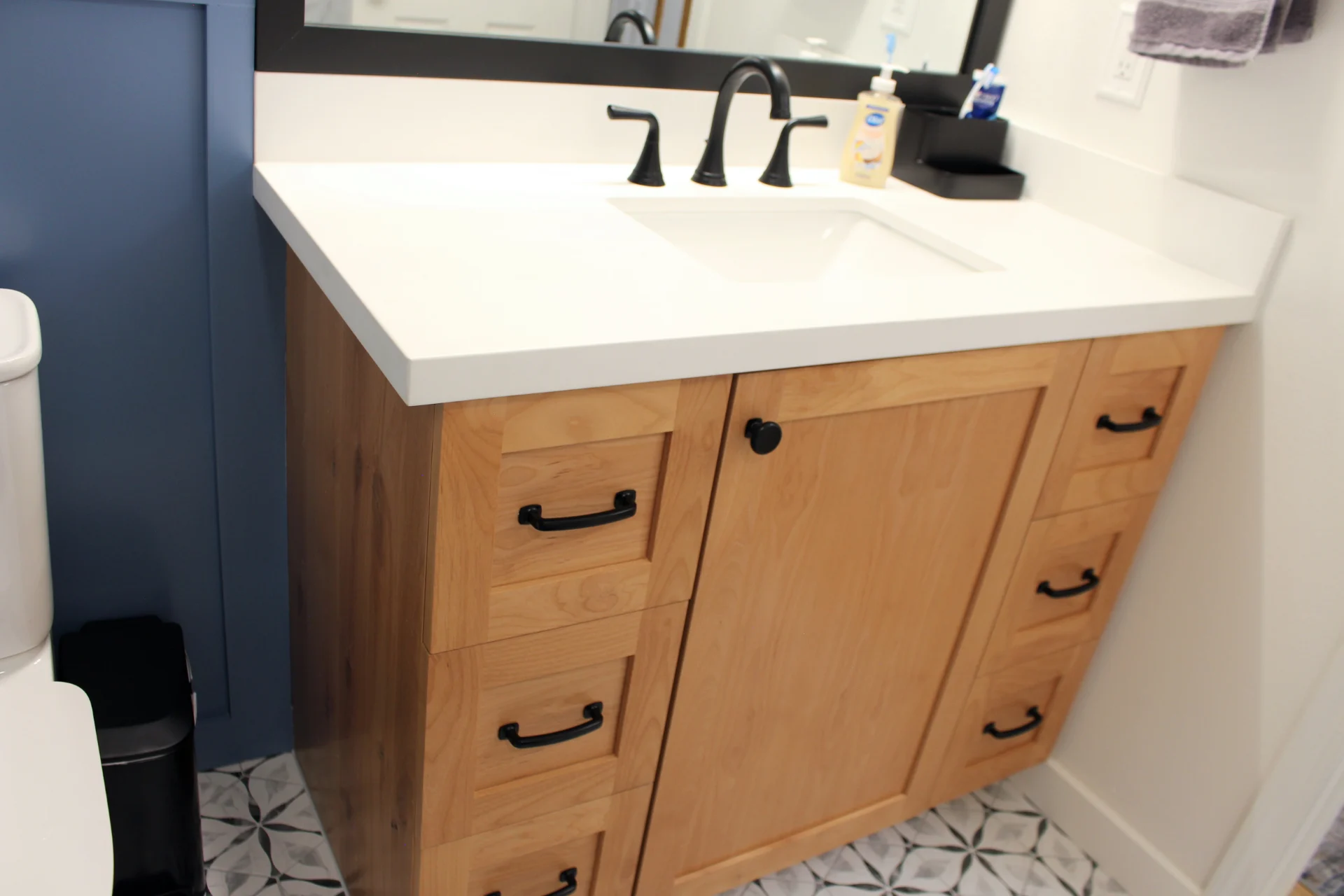 Wooden bathroom vanity with black handles, white countertop, and a black faucet. Geometric tile floor and toiletries add a modern, clean feel.
