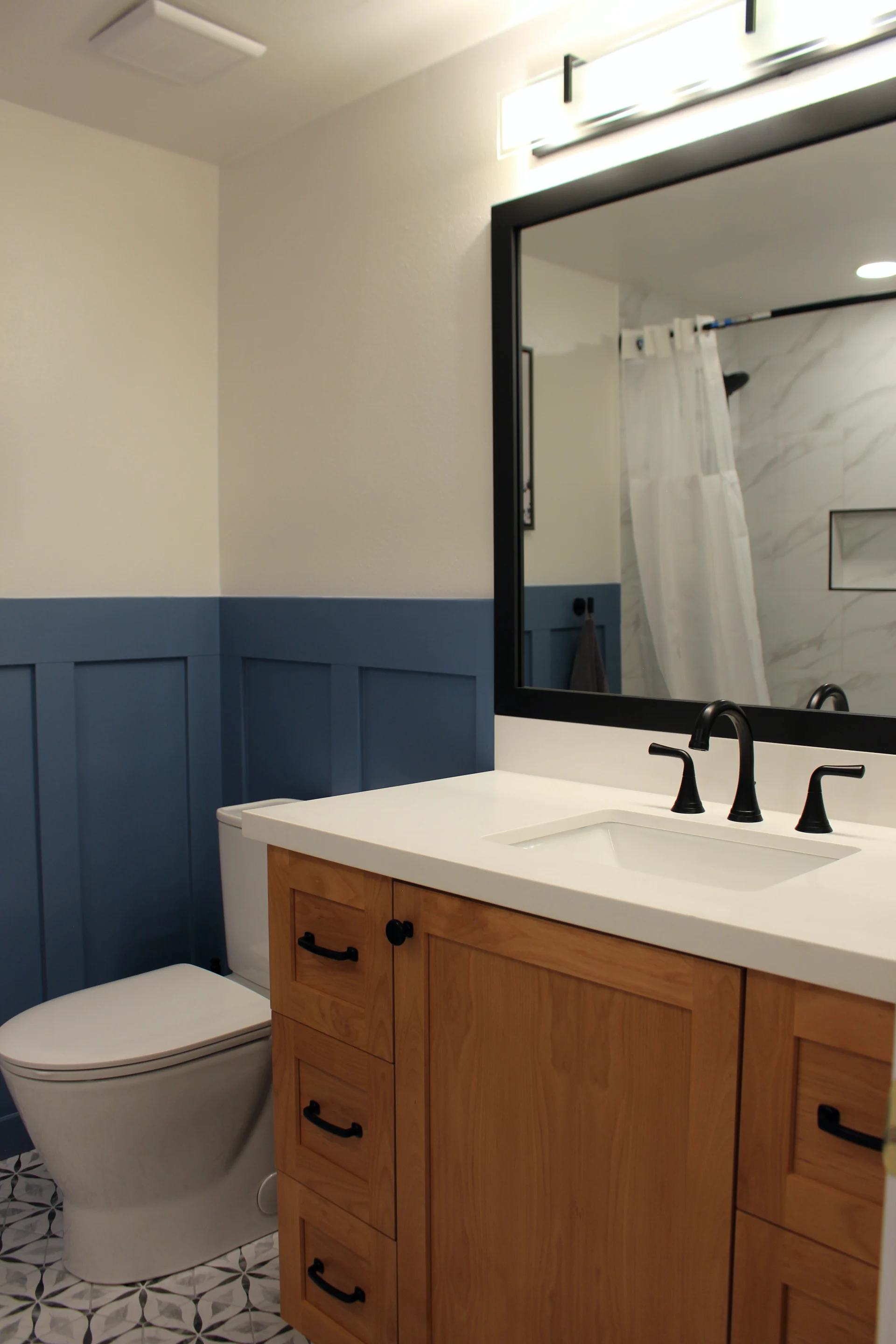 Modern bathroom with wooden vanity, black faucet, and large mirror. Blue wainscoting contrasts with marble shower, creating a sleek, calm ambiance.