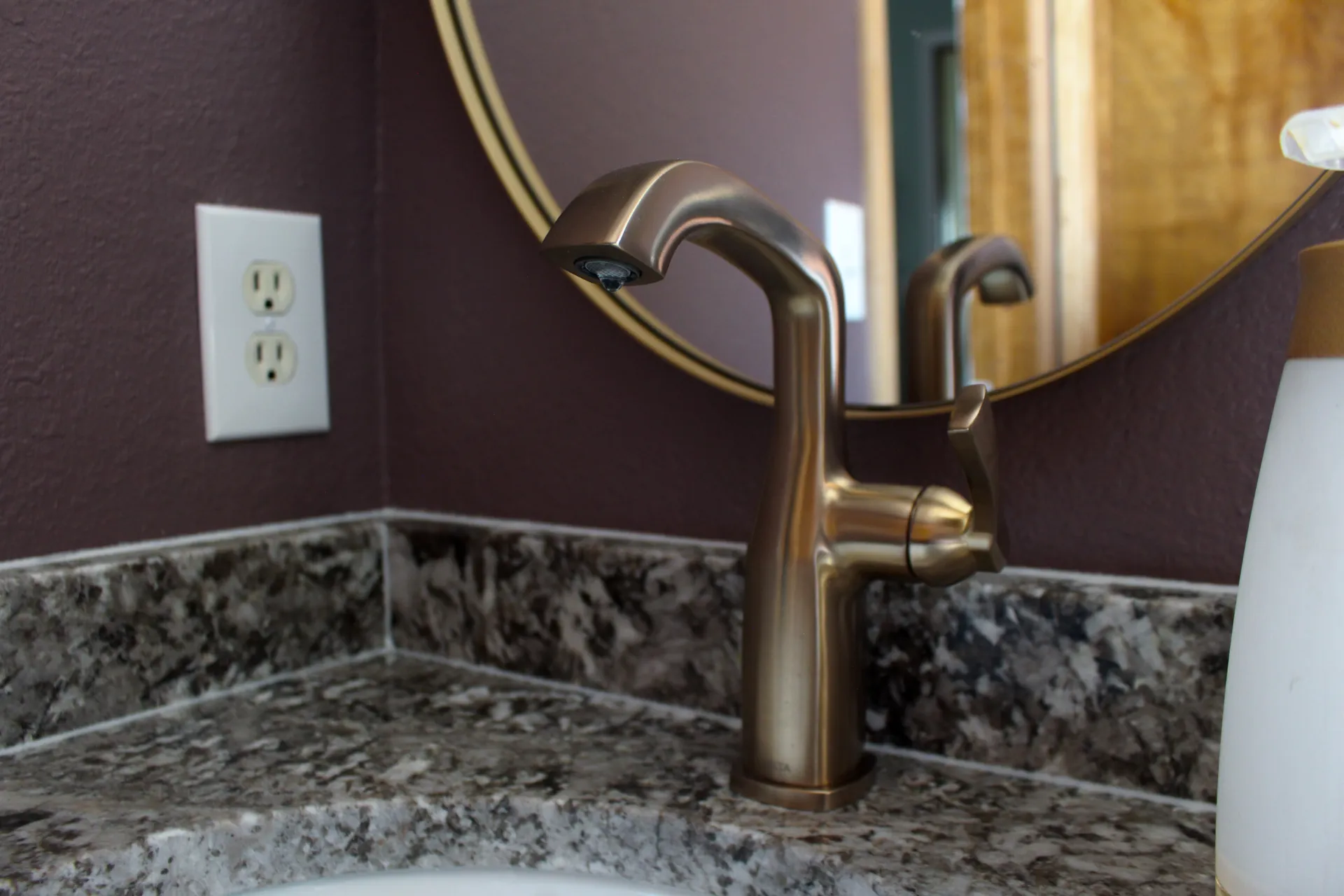 A sleek bronze faucet on a marble countertop with a round mirror in the background. The setting is calm and modern, featuring a deep purple wall.