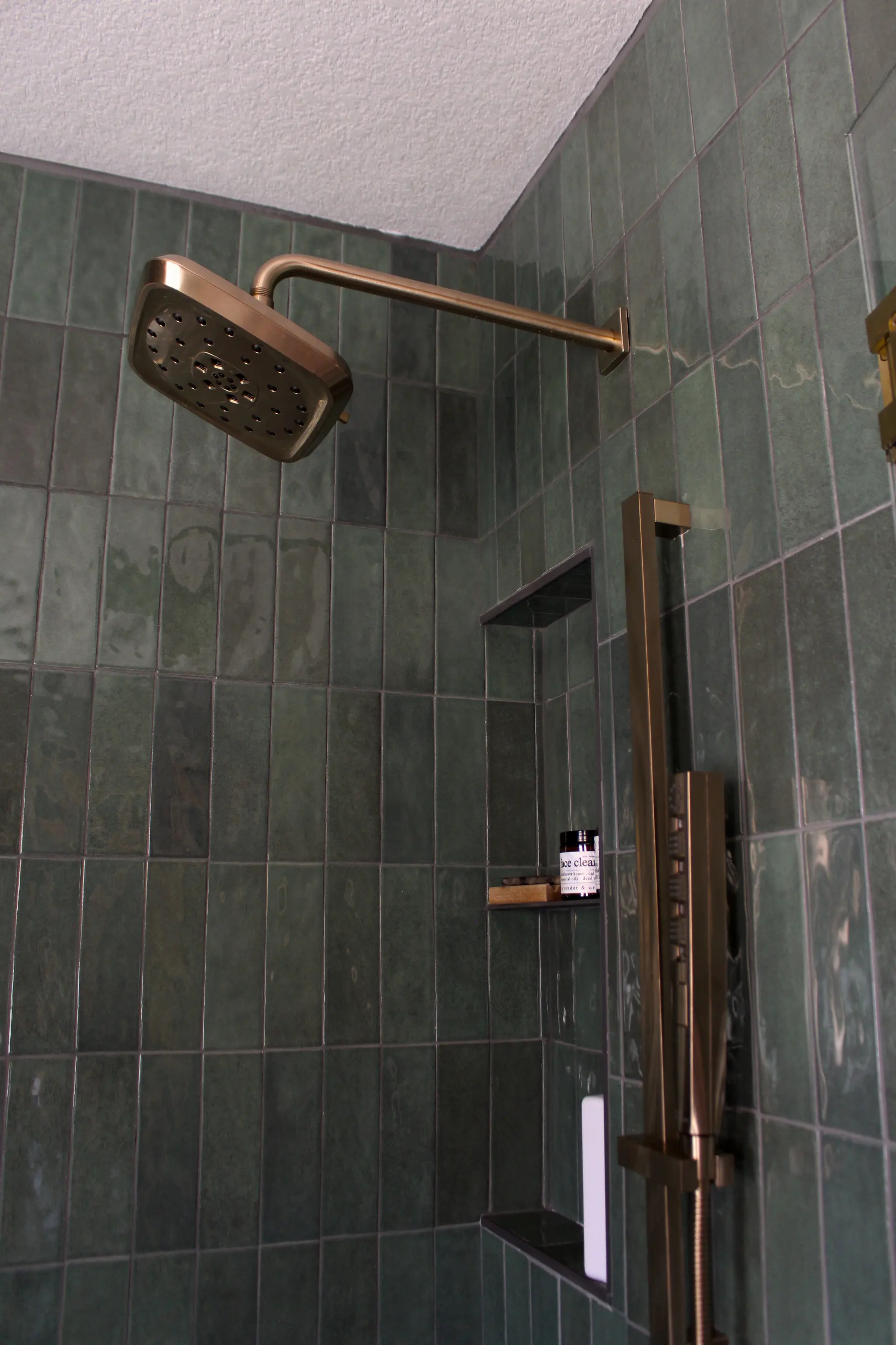 Modern shower with a square, brass showerhead on green tiled walls. A recessed shelf holds shower products, creating a sleek and luxurious feel.