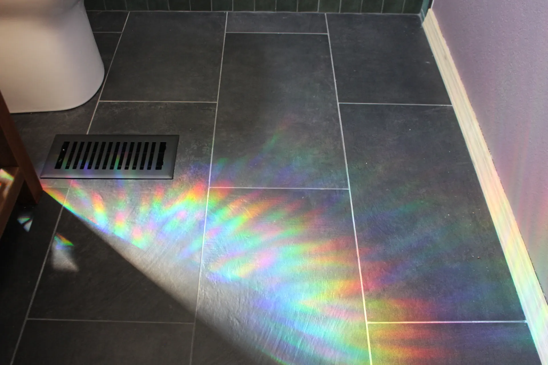 Sunlight casts a vivid rainbow across dark tiled floor next to a vent, creating a colorful, serene atmosphere.