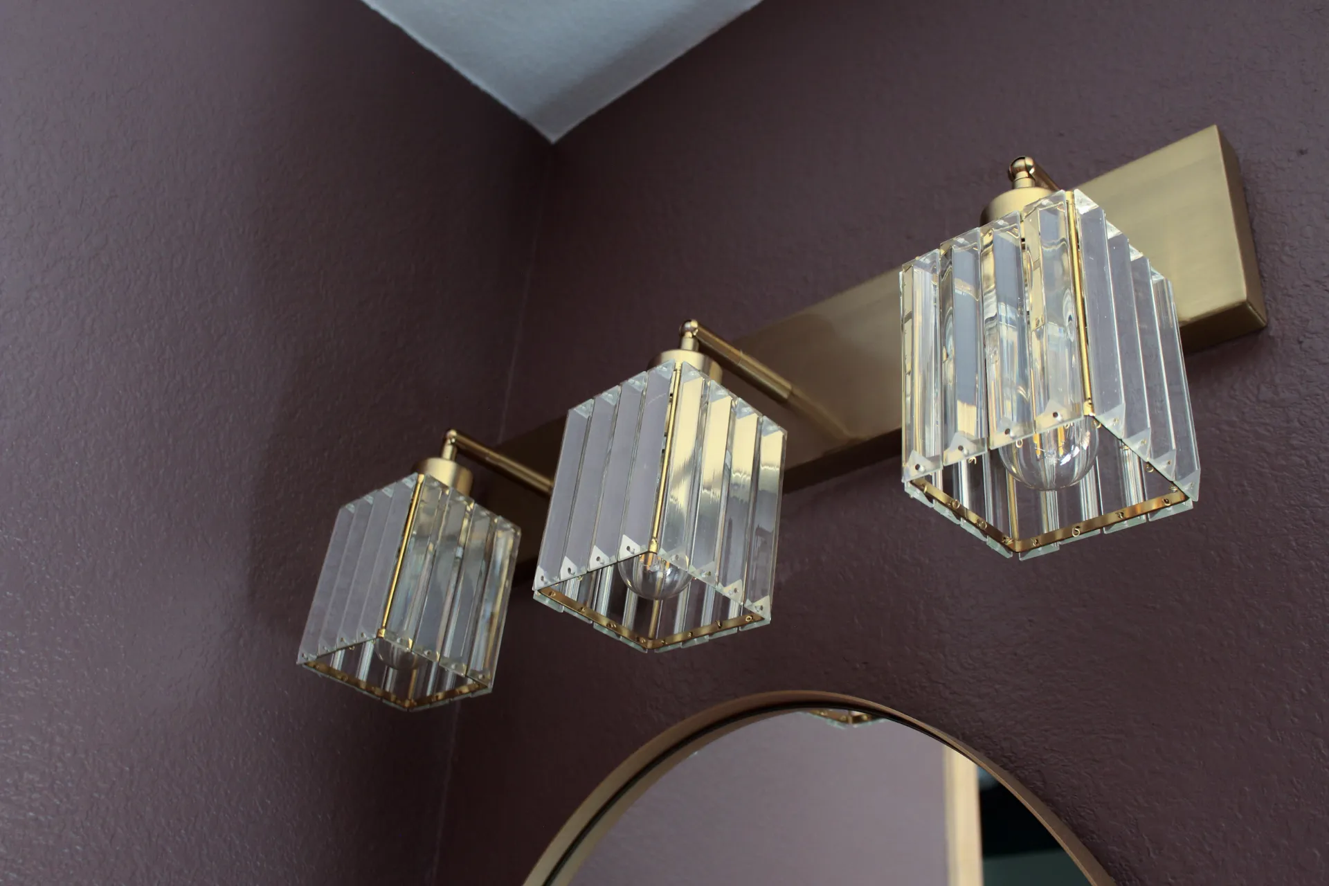 Elegant bathroom light fixture with three gold-accented, faceted glass shades. Mounted on a dark mauve wall, creating a sophisticated ambiance.