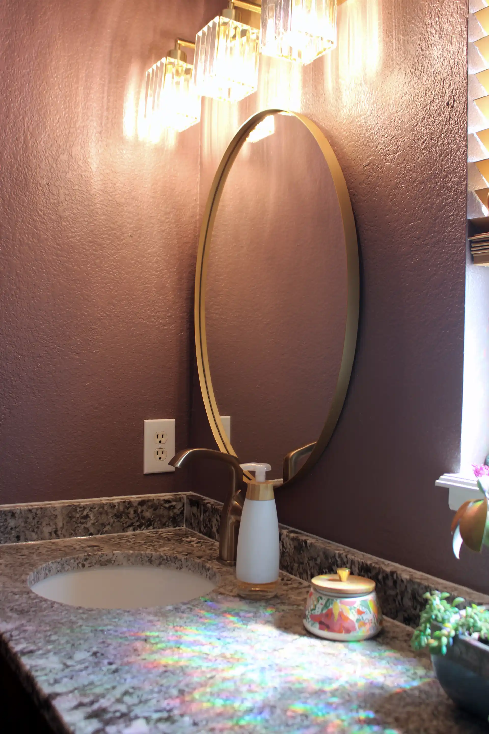 Elegant bathroom corner with a round gold-framed mirror, purple walls, modern gold faucet, granite countertop, and three crystal pendant lights above.