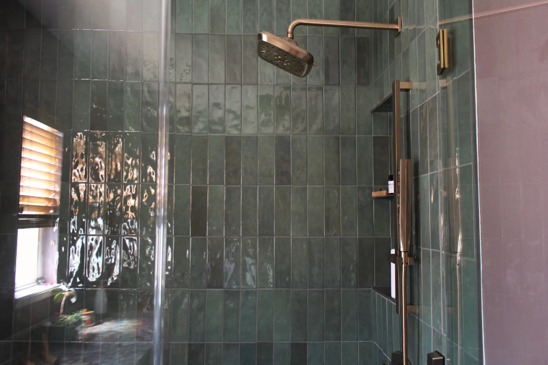 Modern shower with dark green tiles, metal showerhead, and glass door. Natural light streams in from a window with blinds, creating a calm ambiance.