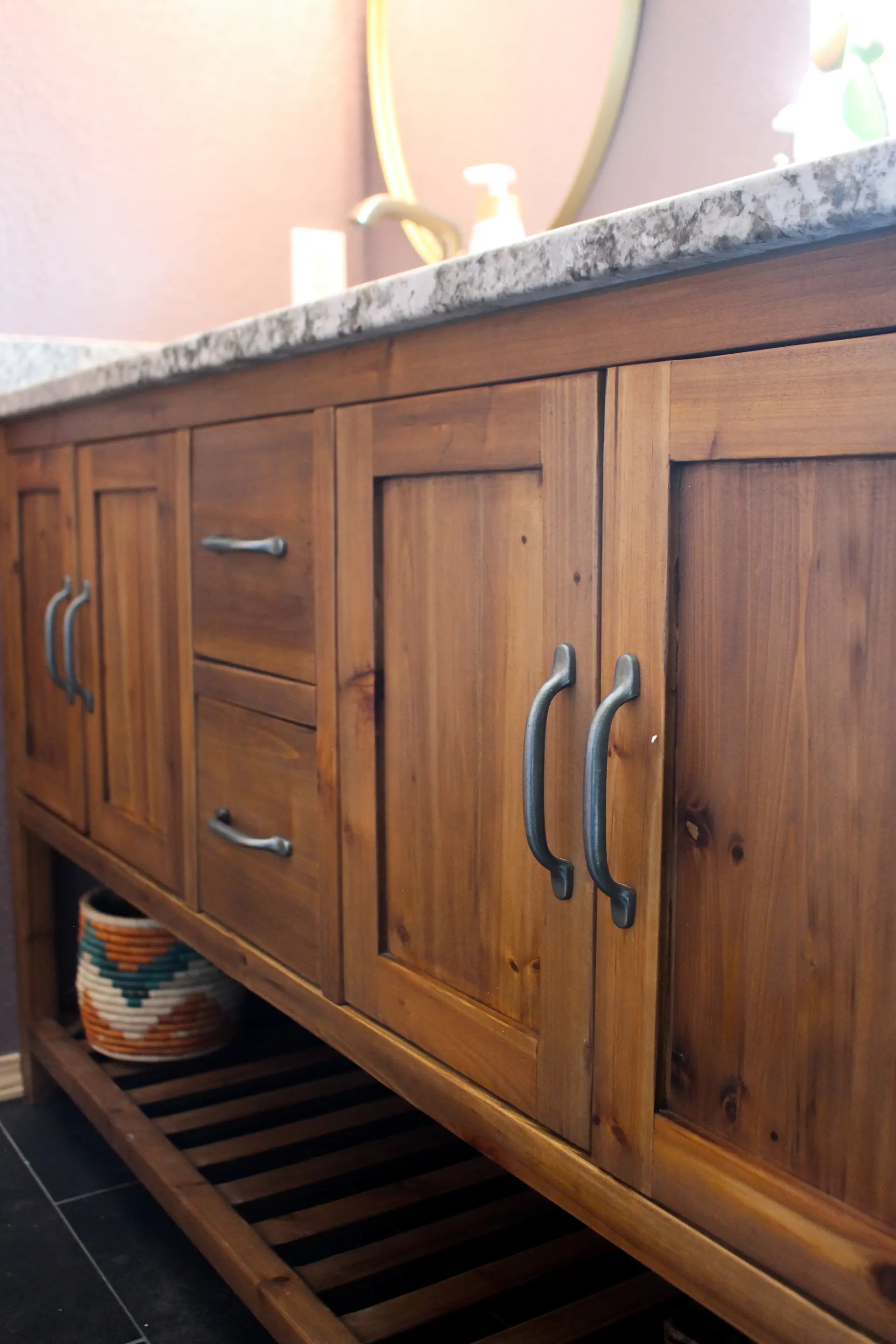 Wooden bathroom vanity with rustic cabinets and metal handles, topped with a speckled marble countertop. A colorful woven basket sits underneath.