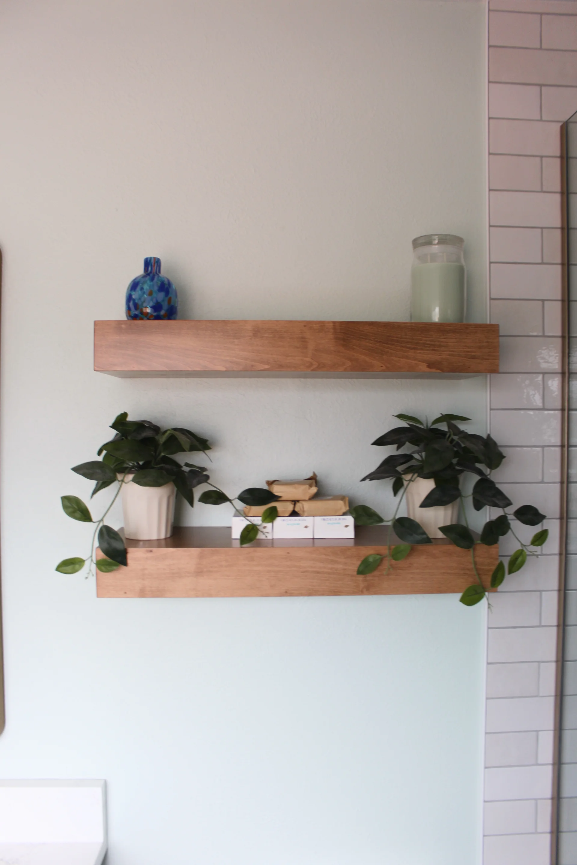 Two wooden floating shelves on a light blue wall; the top shelf holds a blue vase and candle, and the bottom shelf, two potted plants and small boxes.