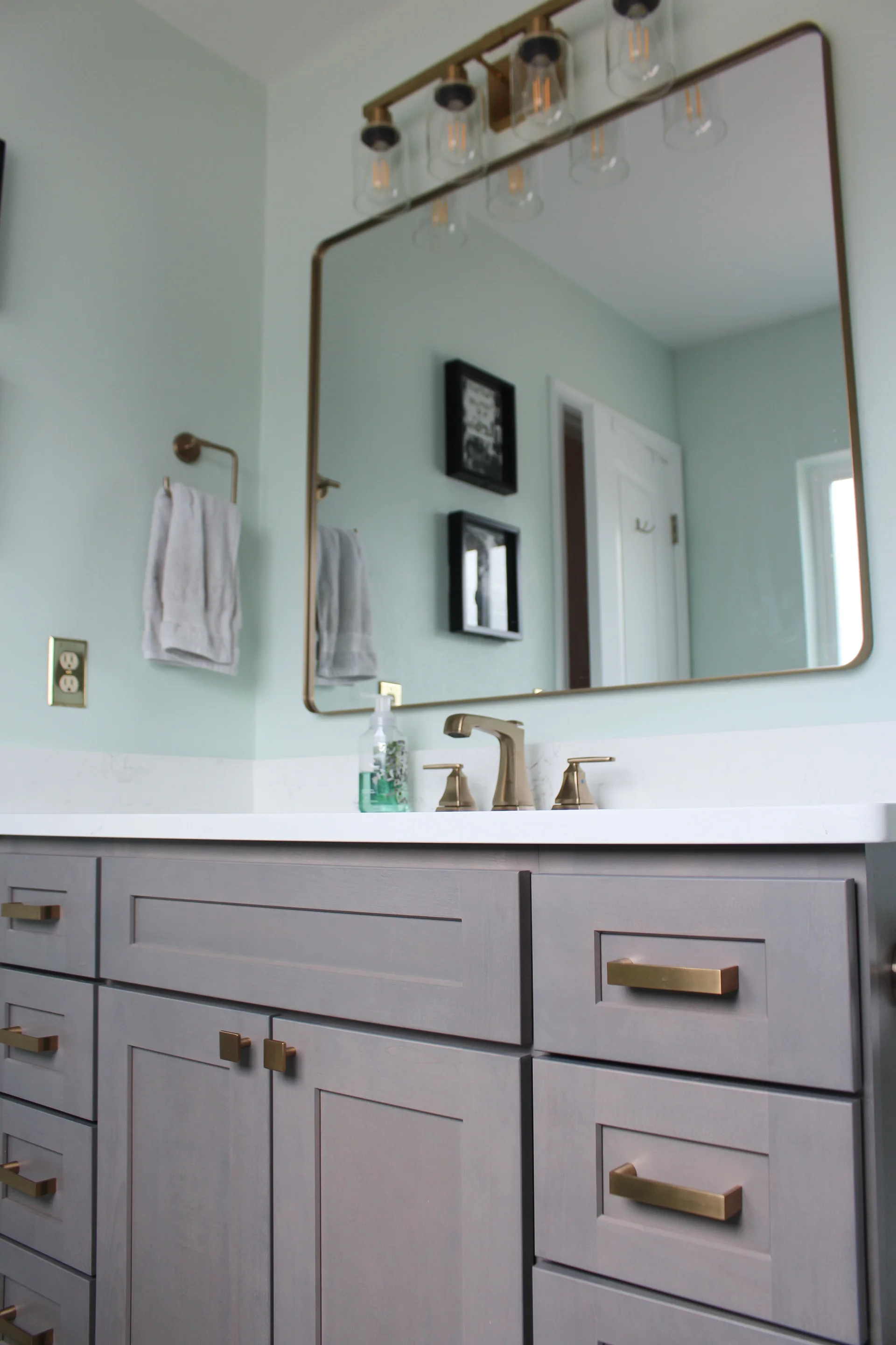 Modern bathroom with light gray cabinets and brass handles. A large mirror, brass faucet, and stylish light fixture enhance the elegant, serene space.