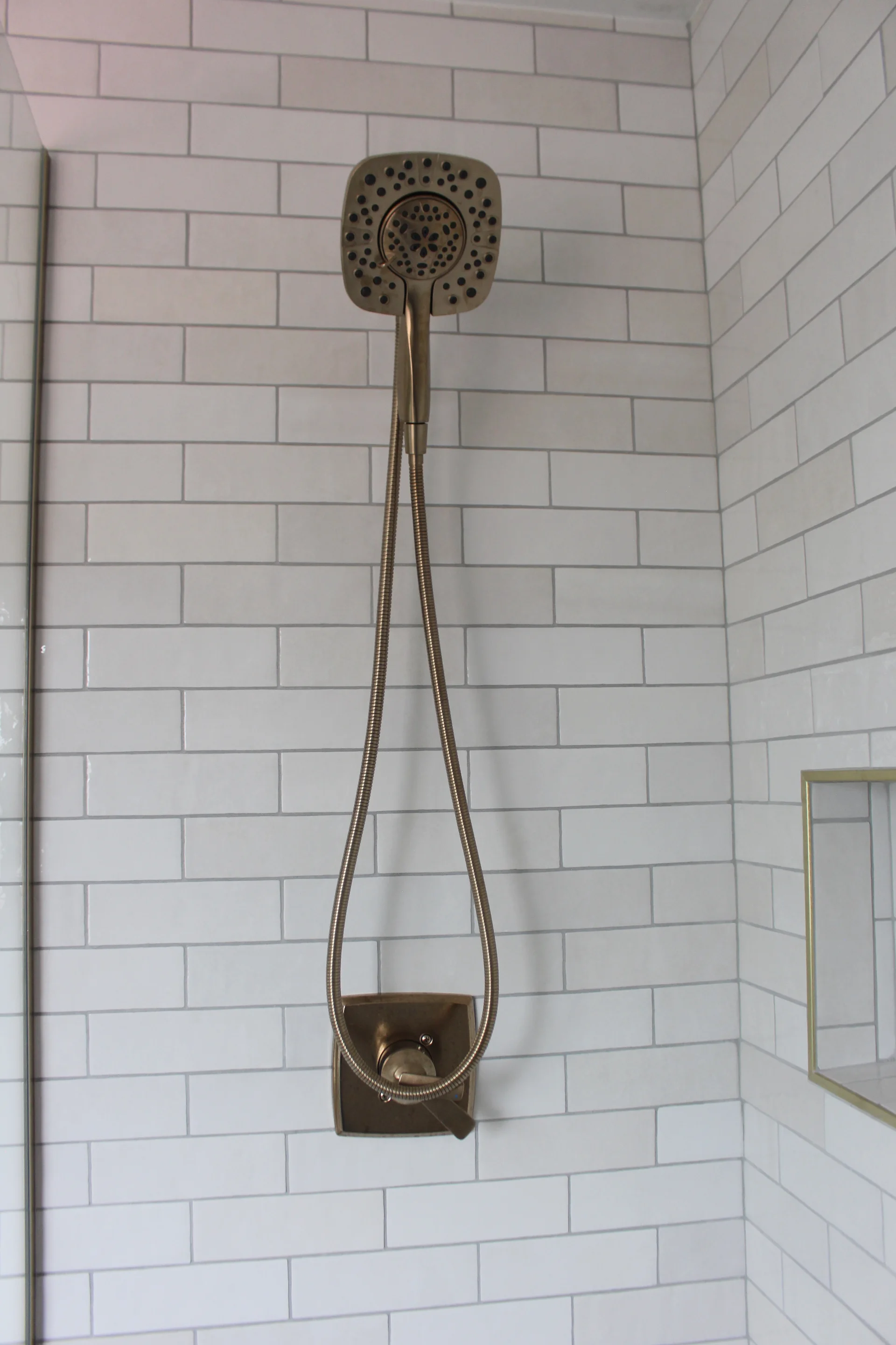 Shower with a square bronze head and hose set against white subway tiles in a bathroom. A small shelf niche is visible to the right.