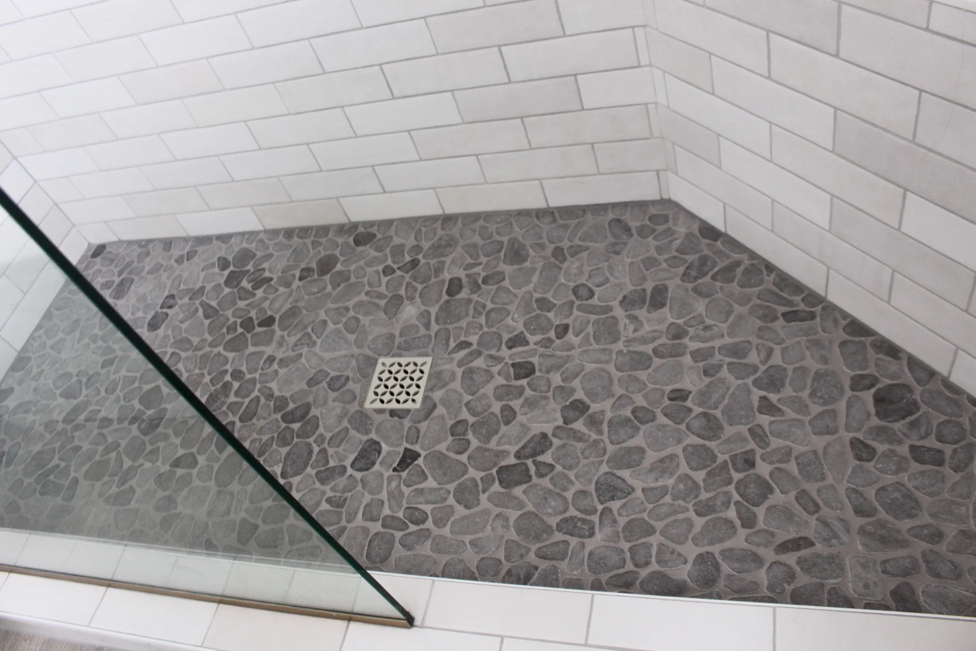 Shower with pebble stone floor, white subway tile walls, and a square pattern drain. A glass panel encloses the modern, clean design.