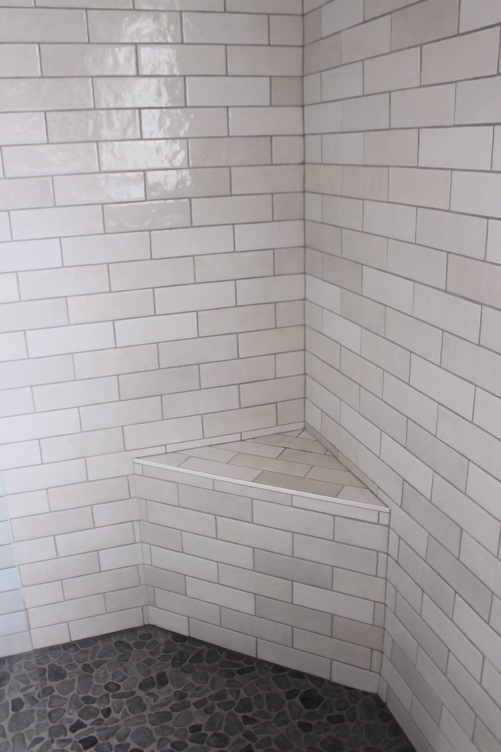 Corner of a shower with white subway tiles and a triangular bench. The floor has a dark pebble-like stone pattern. The scene is clean and modern.