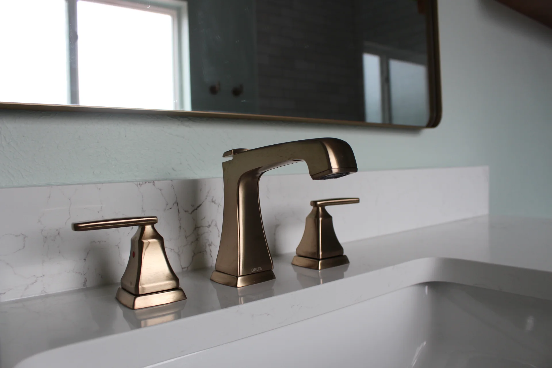 Modern bathroom sink with a sleek bronze faucet and handles on a white marble countertop. A mirror and window are visible in the background.