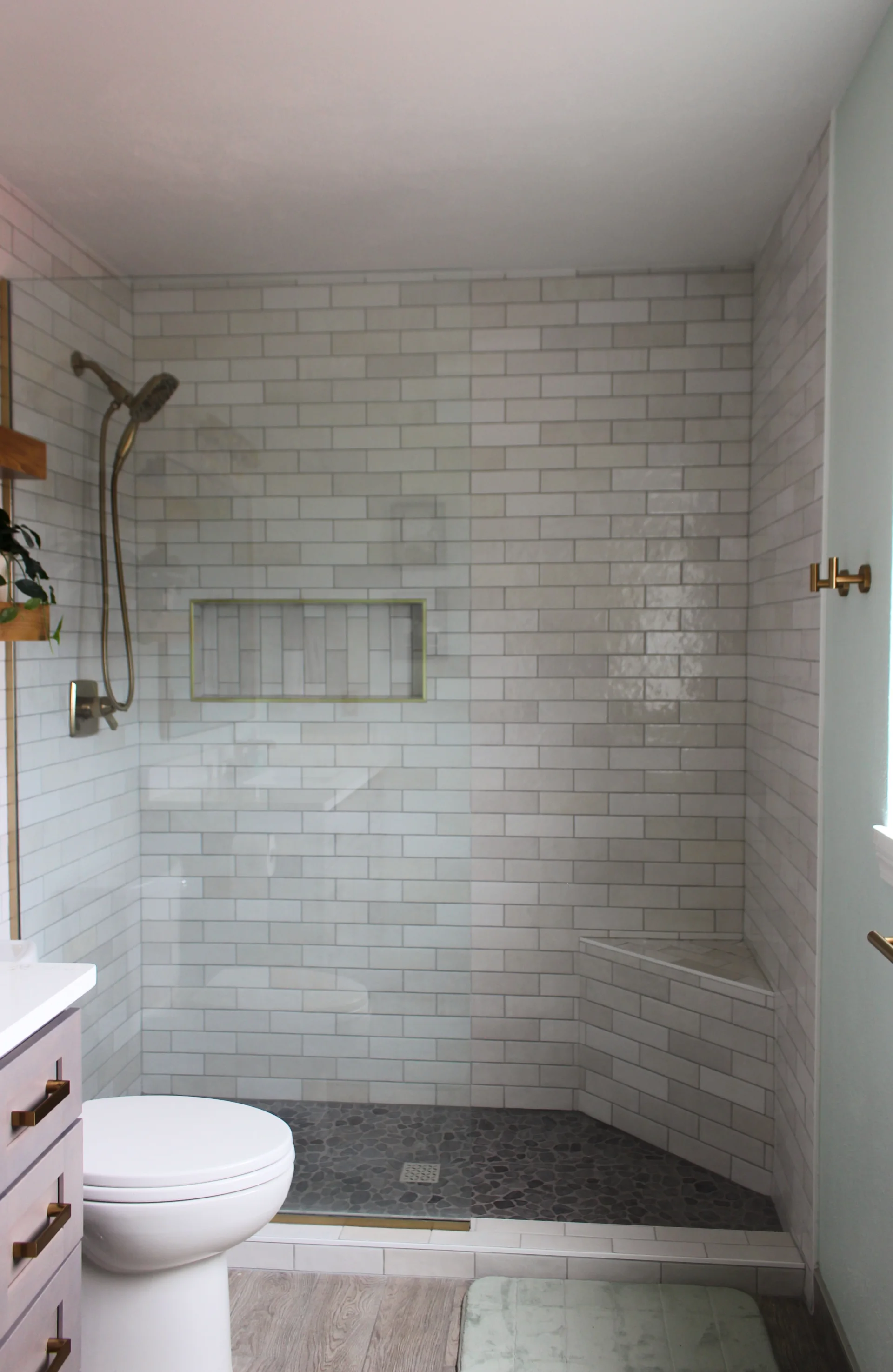 Modern bathroom with a glass-enclosed shower, white subway tiles, and dark pebble flooring. A toilet and vanity with brass handles are visible, creating a clean, minimalist ambiance.