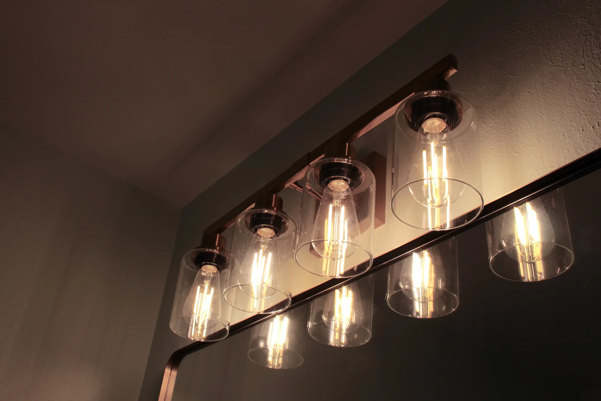 Wall-mounted light fixture with five clear glass shades, each housing a glowing vintage-style bulb. The warm, ambient glow creates a cozy atmosphere.