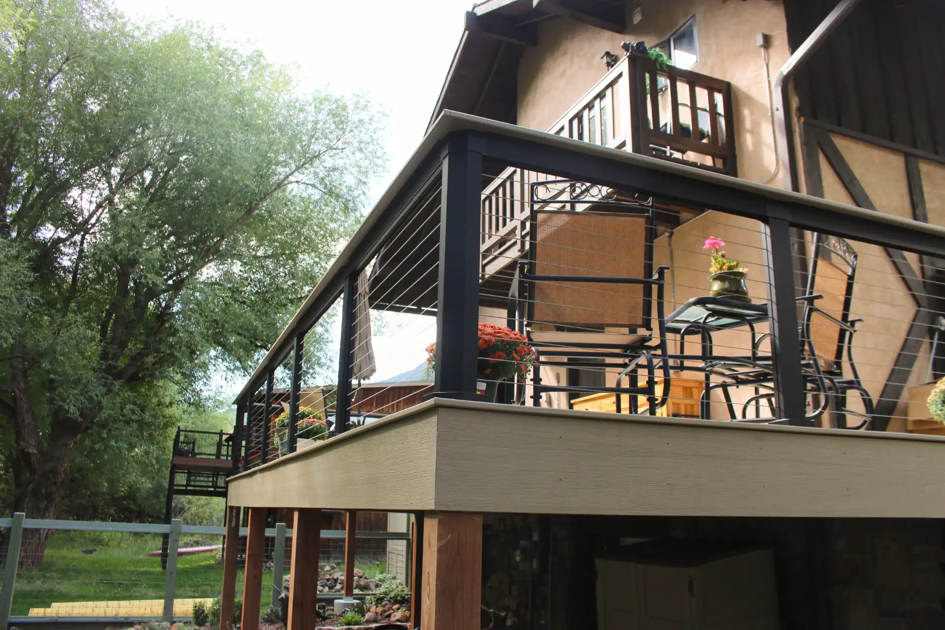 Elevated deck with black railings, outdoor furniture, and potted plants, attached to a rustic house with lush trees in the background. Bright, serene atmosphere.