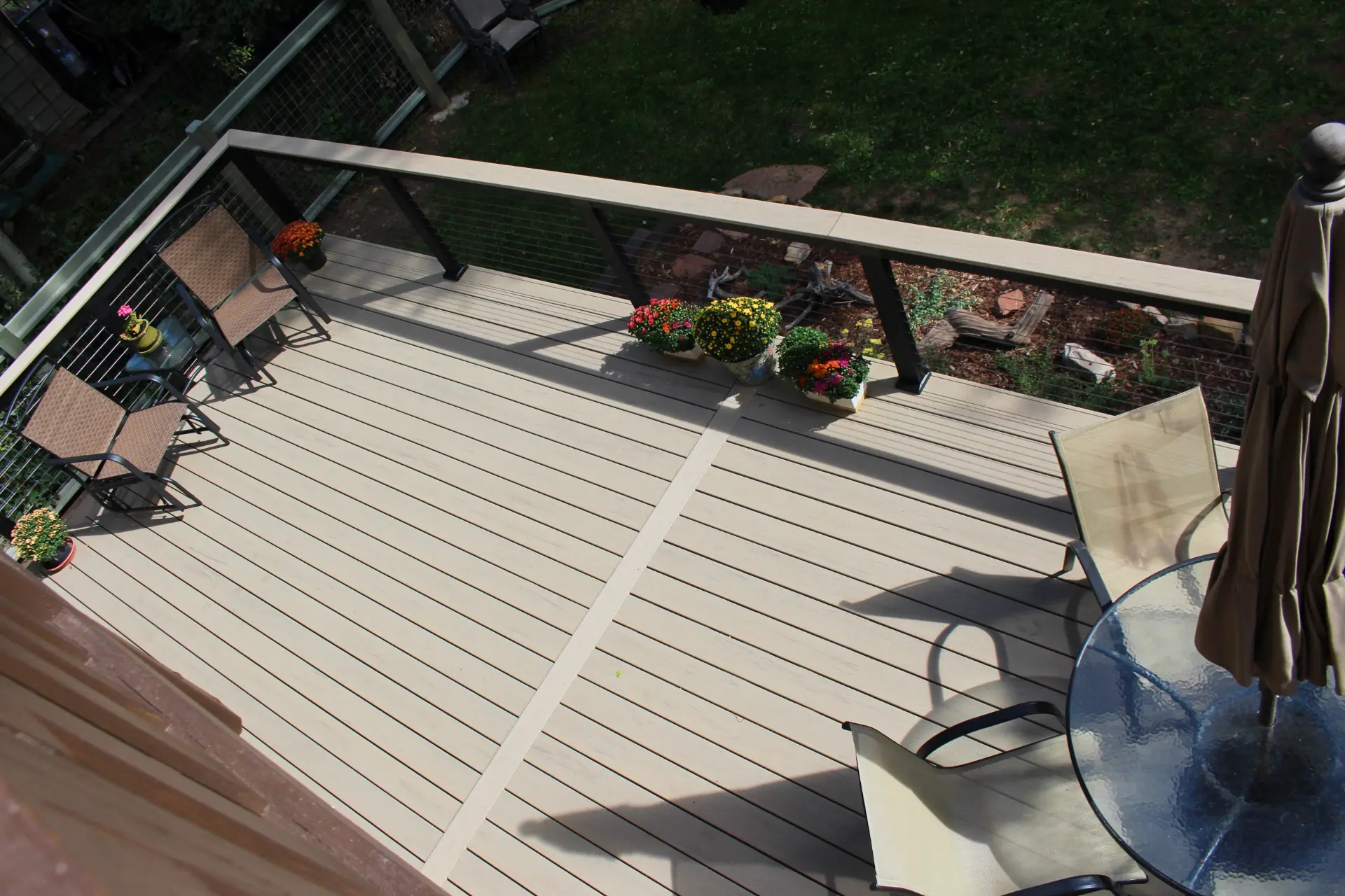 Aerial view of a beige deck with two lounge chairs, colorful potted flowers, and a glass table with closed umbrella. The setting feels peaceful and inviting.