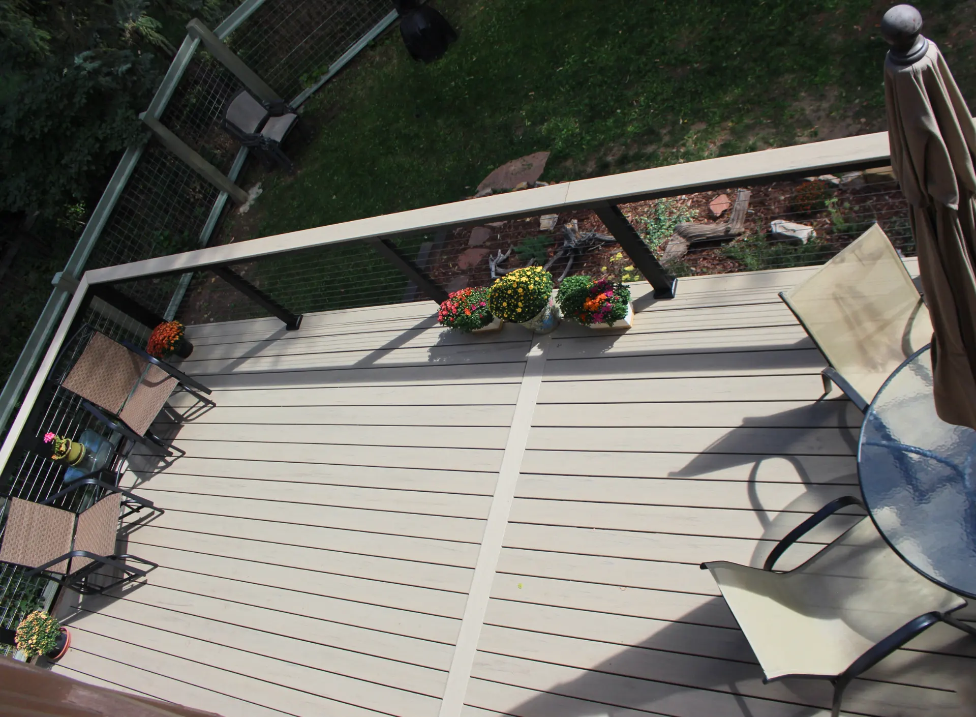 A beige deck with potted flowers, two chairs, and a glass table with an umbrella. The setting overlooks a grassy yard, creating a cozy outdoor space.