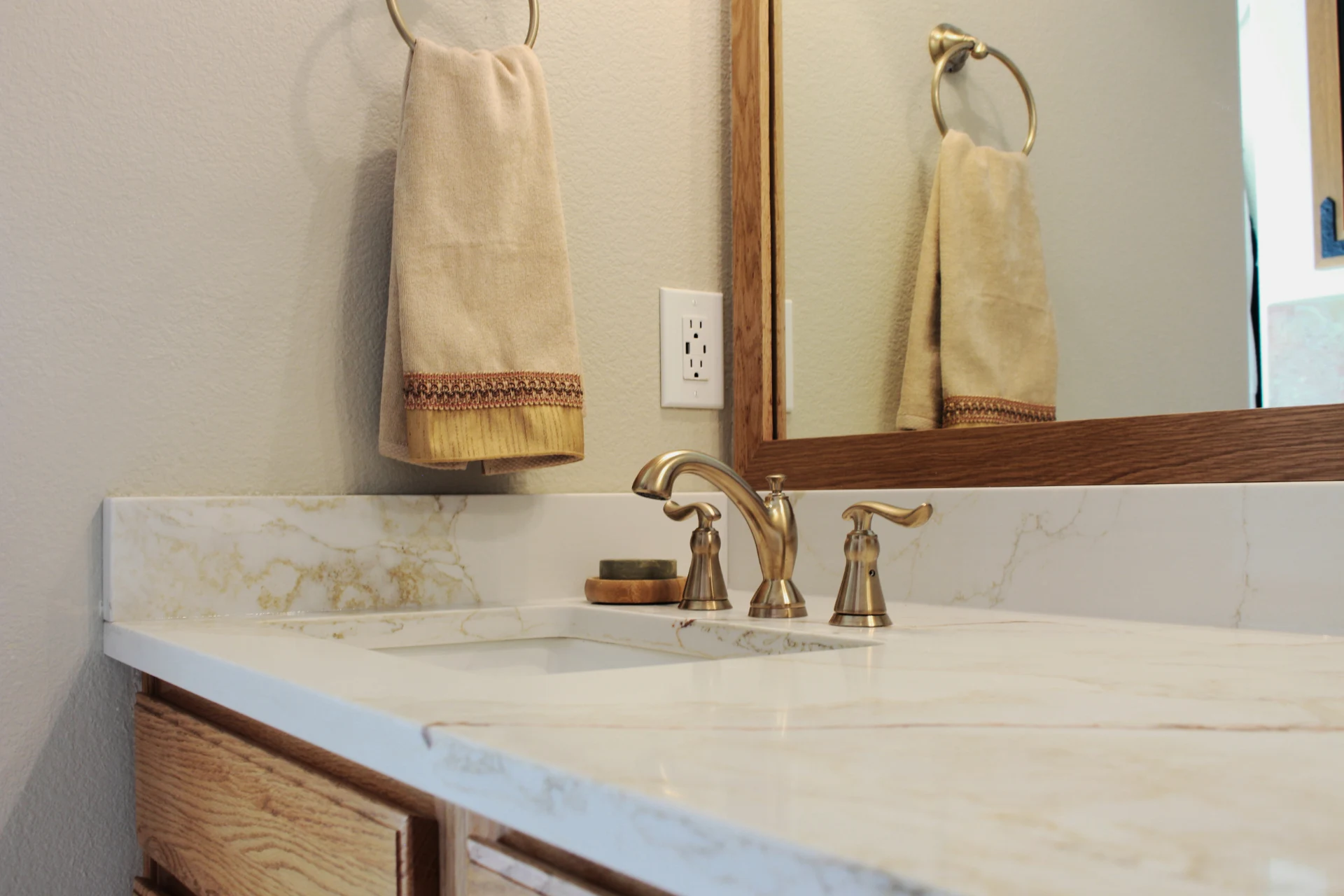 Elegant bathroom vanity with a marble countertop, brass faucet, and plush beige towel hanging. Framed mirror reflects light, creating a warm atmosphere.