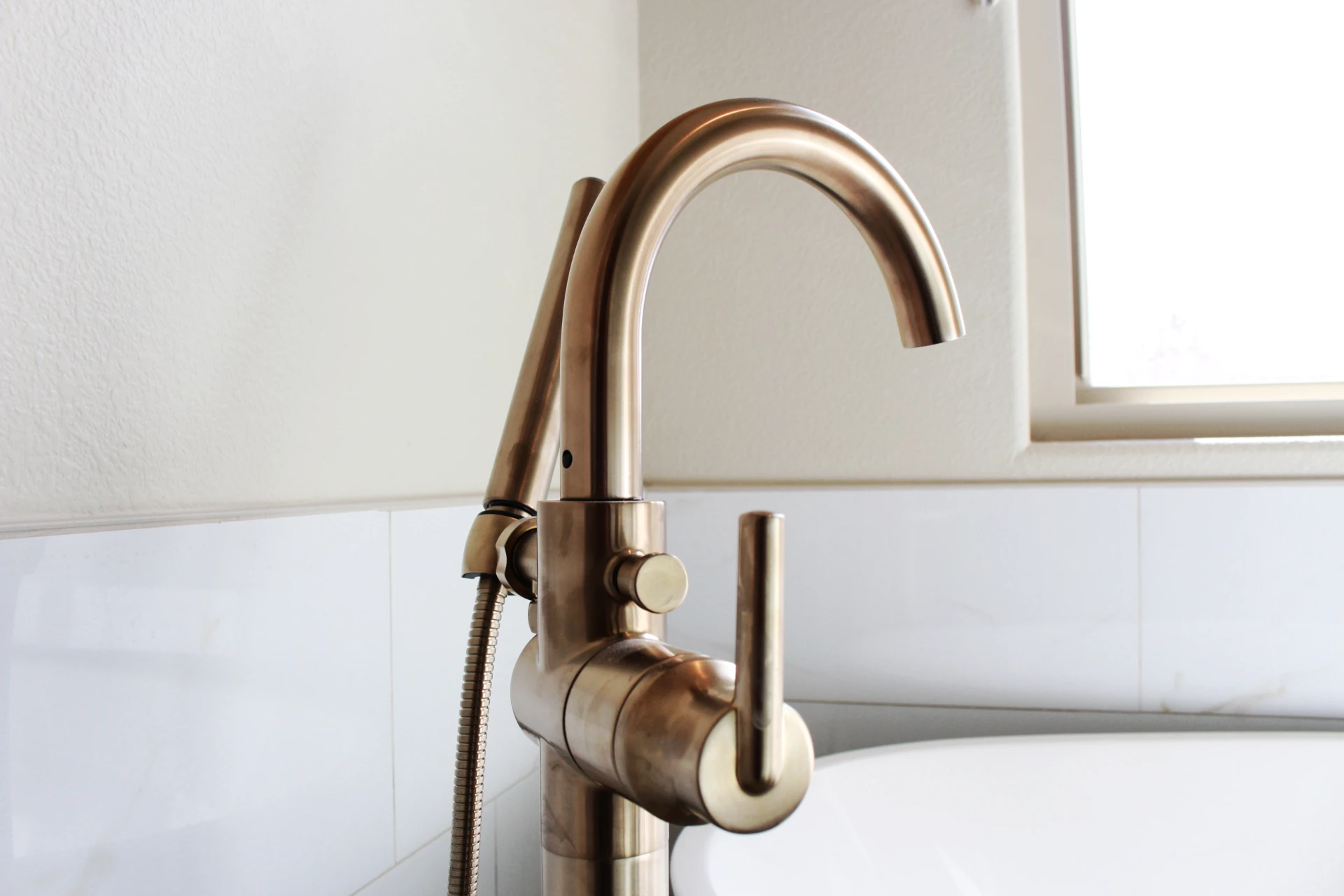 Elegant brass bathroom faucet with a sleek, curved spout is mounted beside a white bathtub. Soft natural light filters through a nearby window.