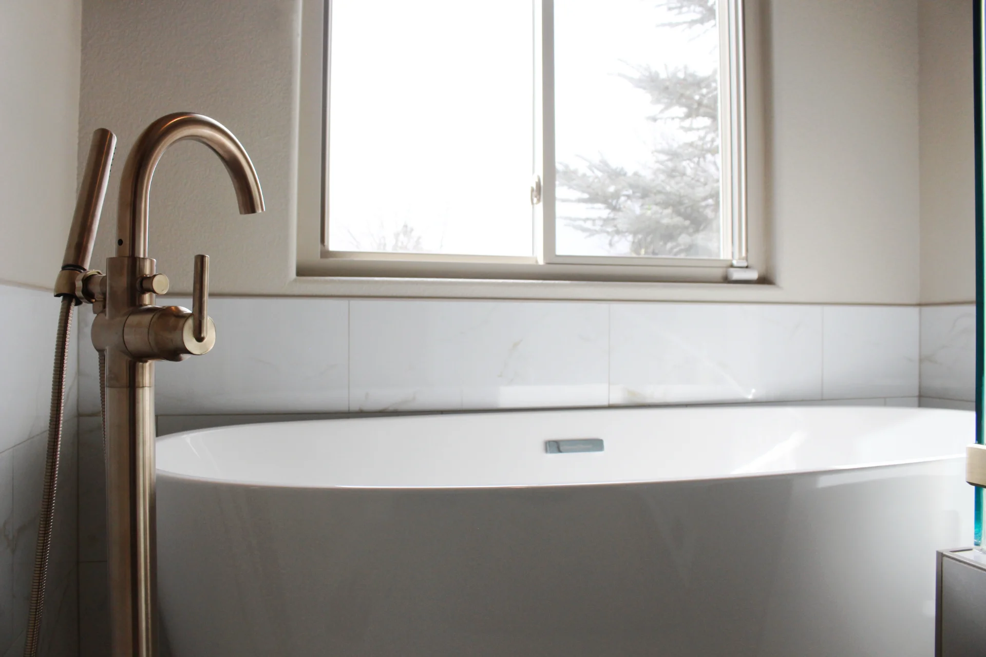Modern bathroom with a freestanding white bathtub and elegant gold faucet. A window above the tub lets in soft natural light, creating a serene atmosphere.