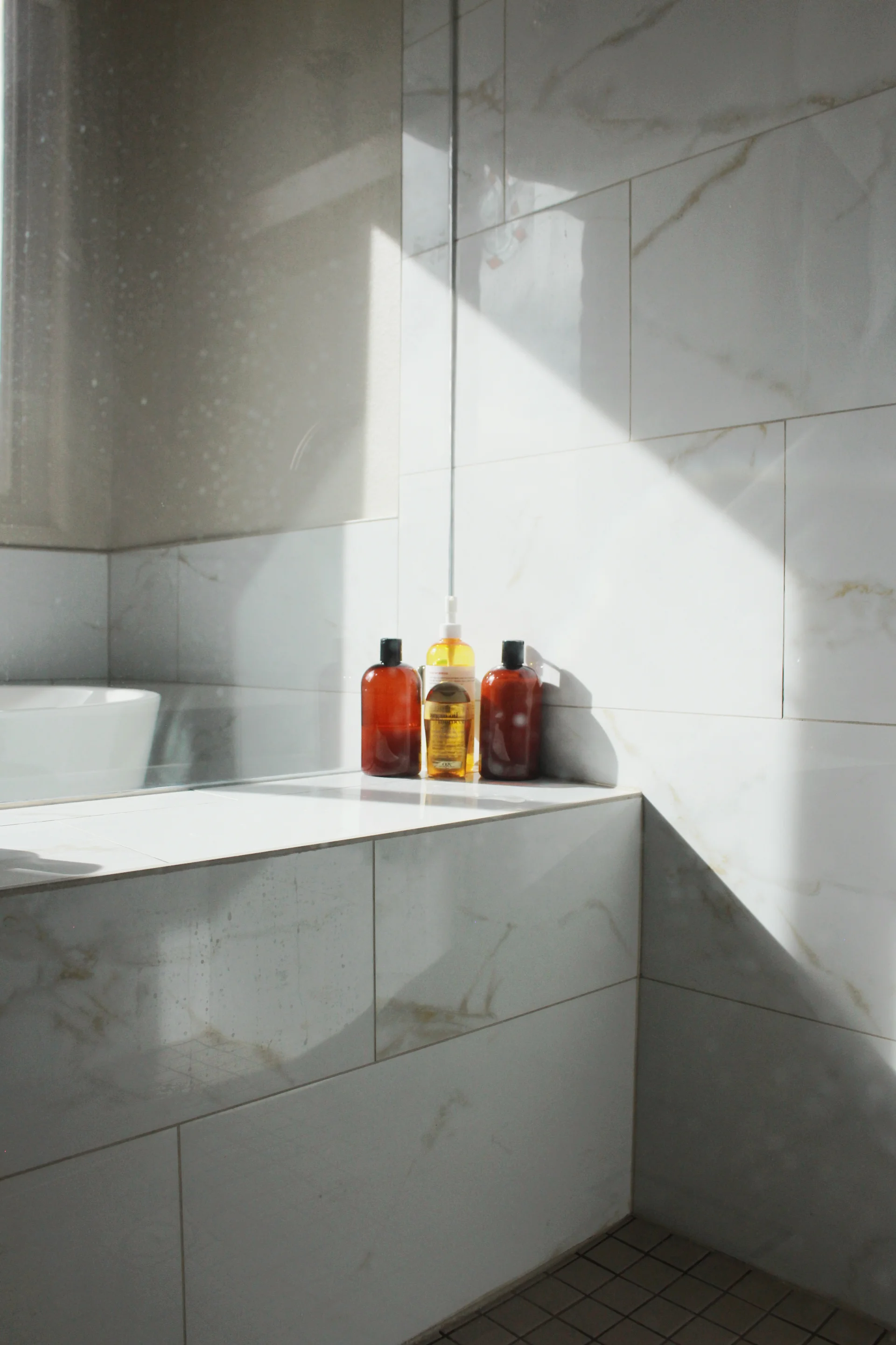 Sunlight streams into a white marble-tiled bathroom, highlighting three amber and clear bottles on the ledge, creating a serene and clean ambiance.