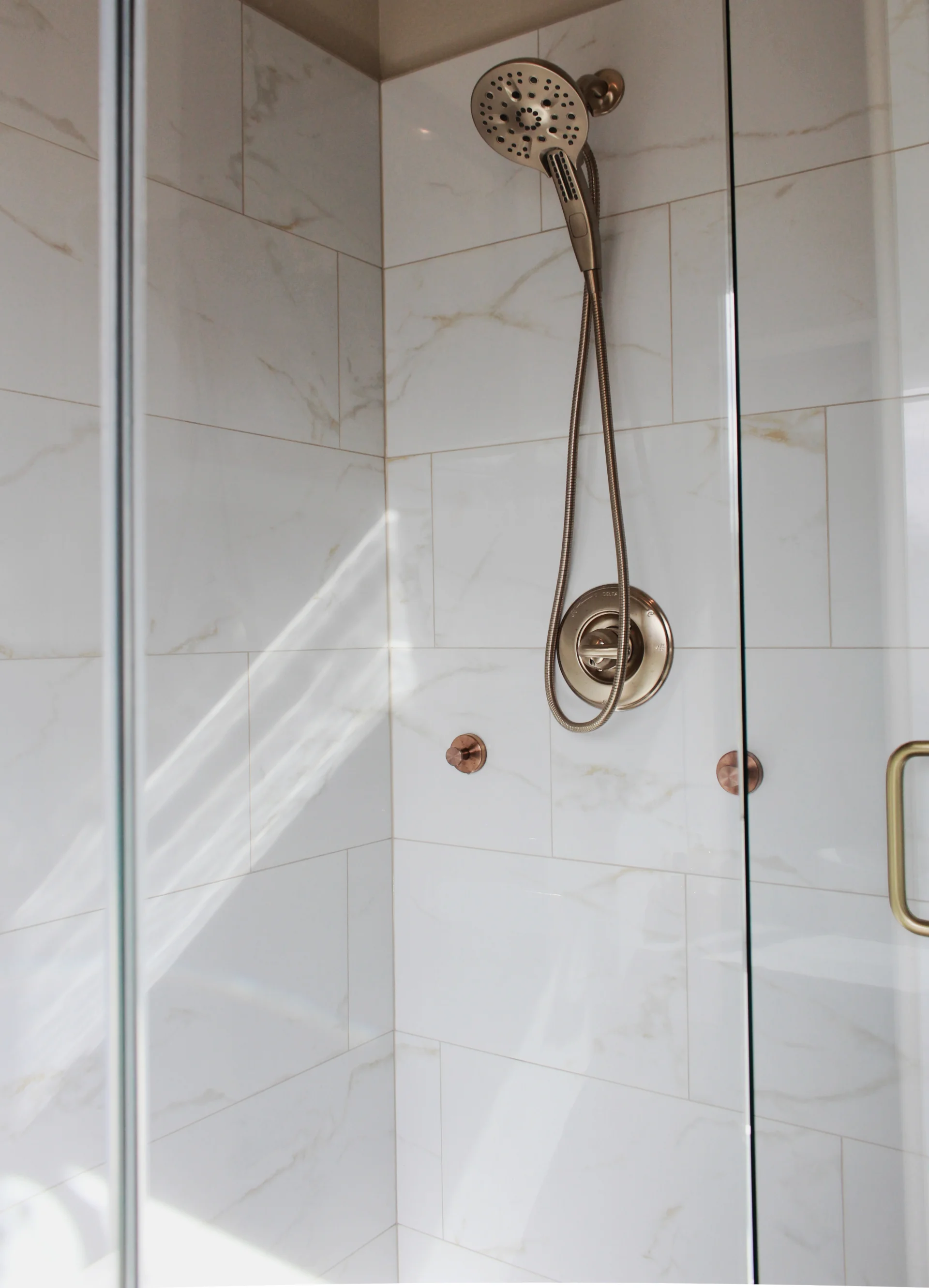 A modern shower with a sleek chrome showerhead and hose is set against white marble tiles. Sunlight streams in, creating a bright, clean atmosphere.
