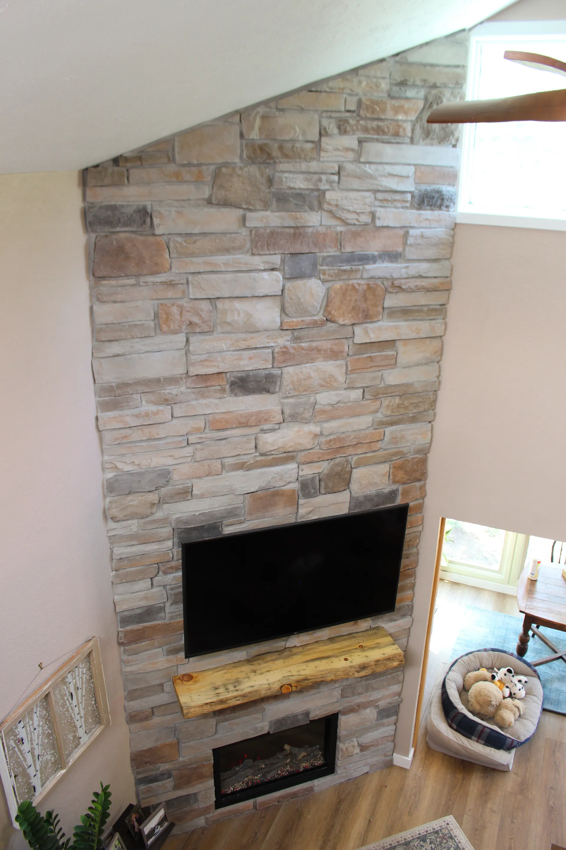 Stone fireplace wall with mounted TV and wooden mantel. Below is an electric fireplace. Cozy dog bed with toys near a bright window, wood floors.