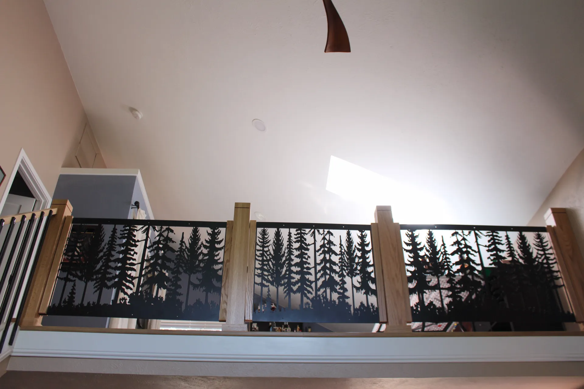 A loft railing with a woodland silhouette design features tall trees between wooden posts. Soft light from a skylight creates a calm, serene atmosphere.