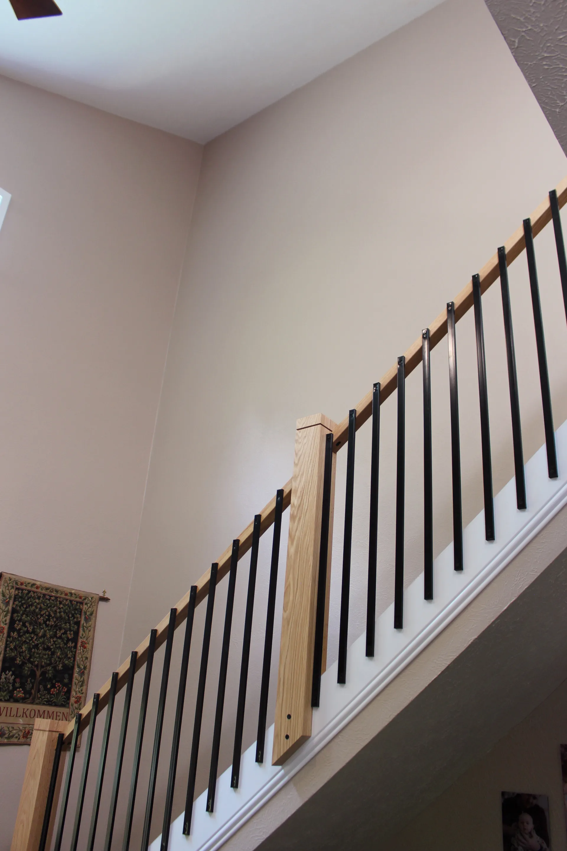 View of a staircase with a modern railing featuring vertical black metal balusters and light wood supports. The beige walls create a calm, spacious atmosphere. A tapestry is partially visible on the lower left wall.