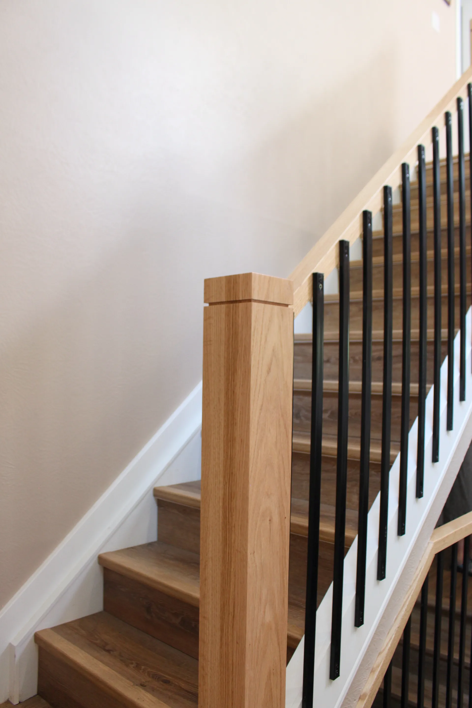 Staircase with wooden steps and a light wood newel post. Black metal balusters contrast with a white wall, creating a modern, minimalist look.