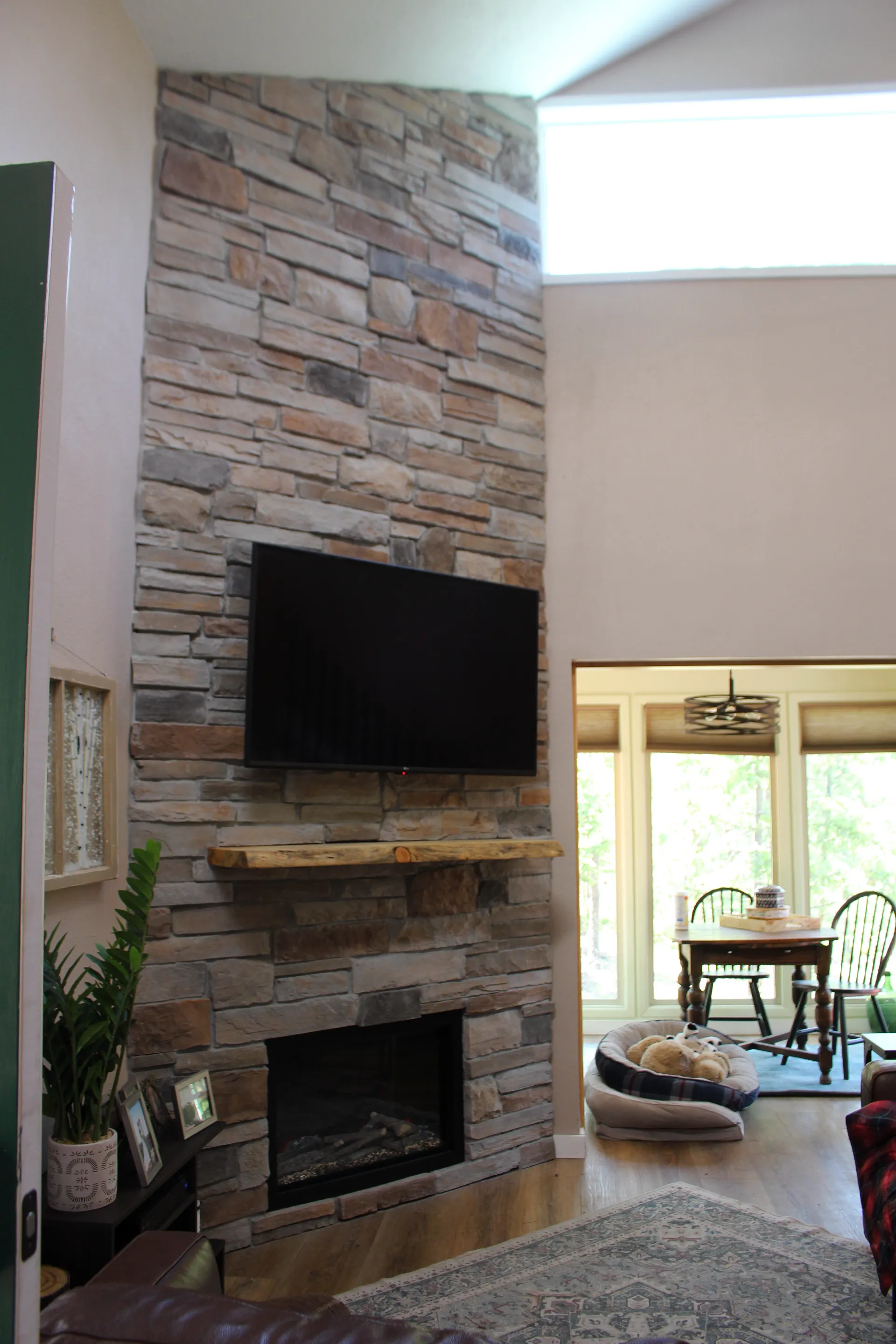 A modern living room features a stone fireplace with a mounted TV above. A dining area with natural light is visible through an archway, creating a cozy ambiance.