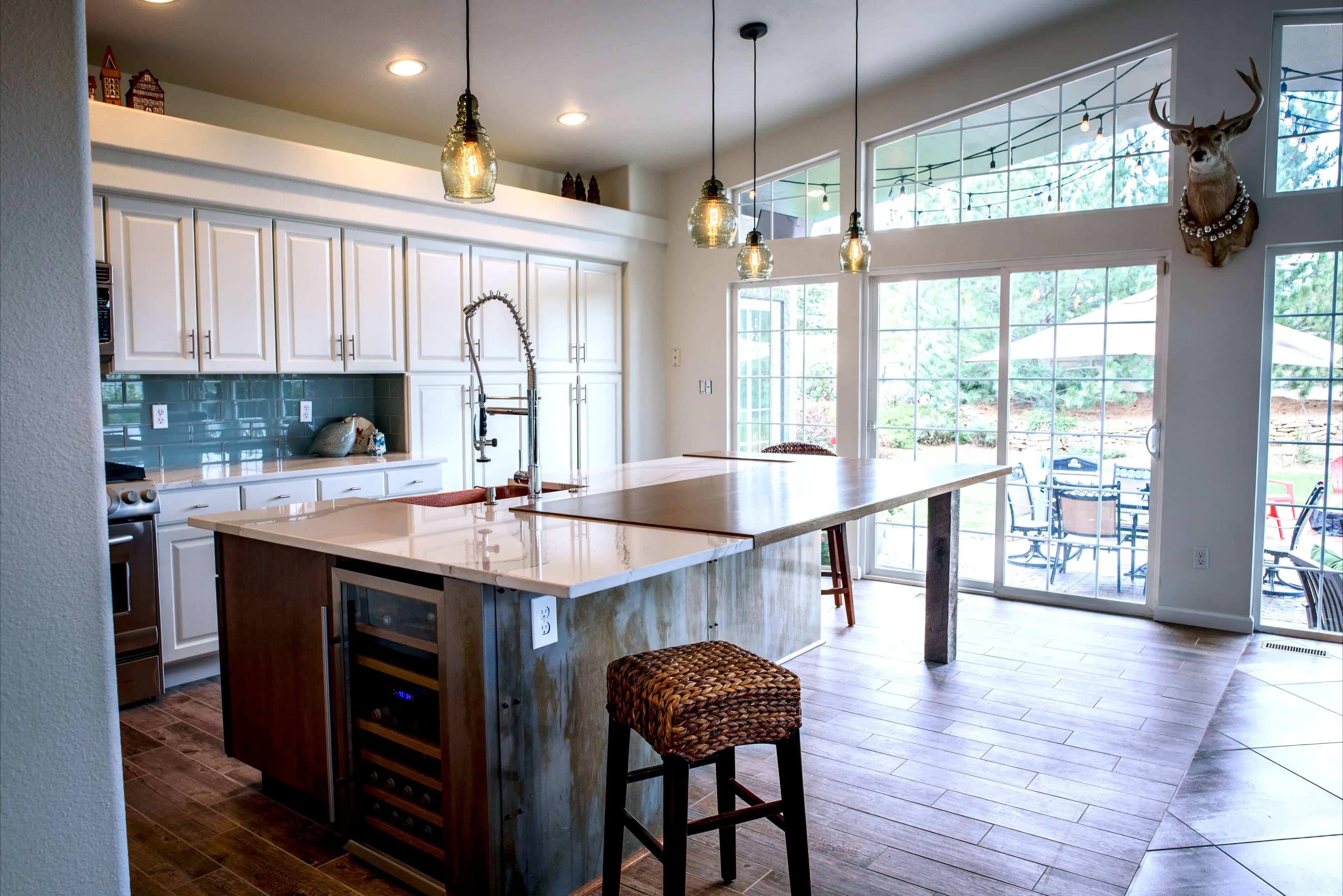 A remodeled kitchen with a blue tiled splash wall, wooden floors, a hammered copper sink, white cabinets, and a wooden breakfast bar and custom hanging lighting