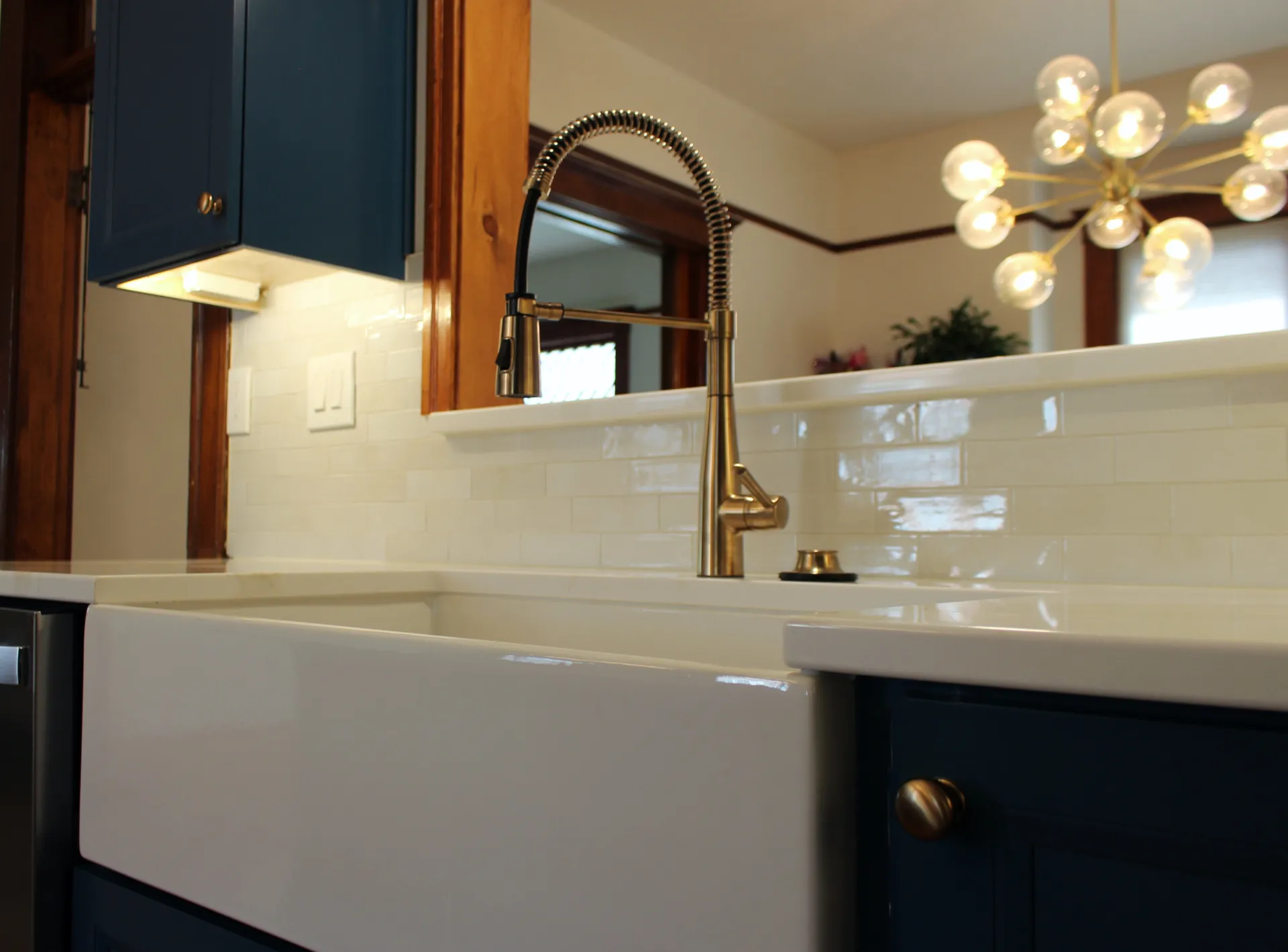 Modern kitchen with gold faucet, white farmhouse sink, and deep blue cabinets. White subway tiles and a chic chandelier create a stylish ambiance.