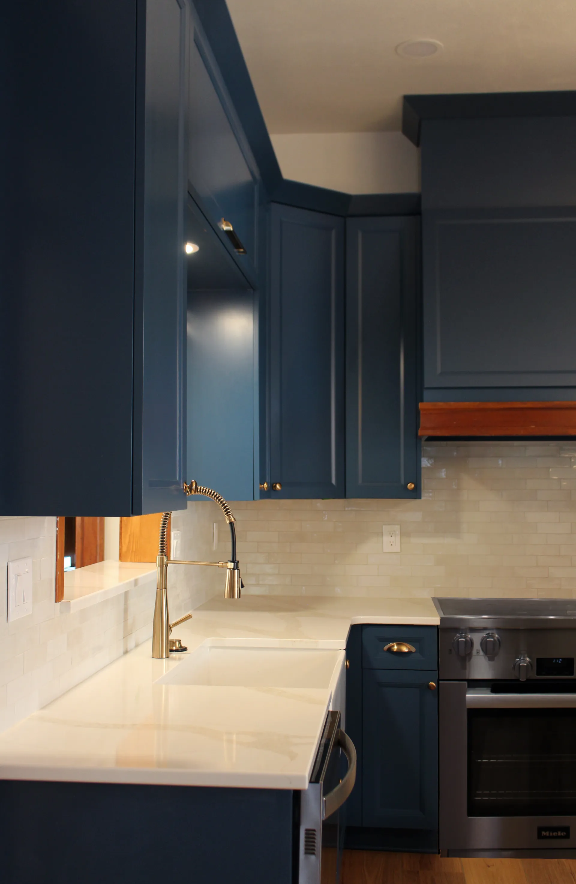 Modern kitchen with blue cabinets and a gold faucet over a white countertop. Stainless steel oven, beige tiled backsplash, and warm lighting.